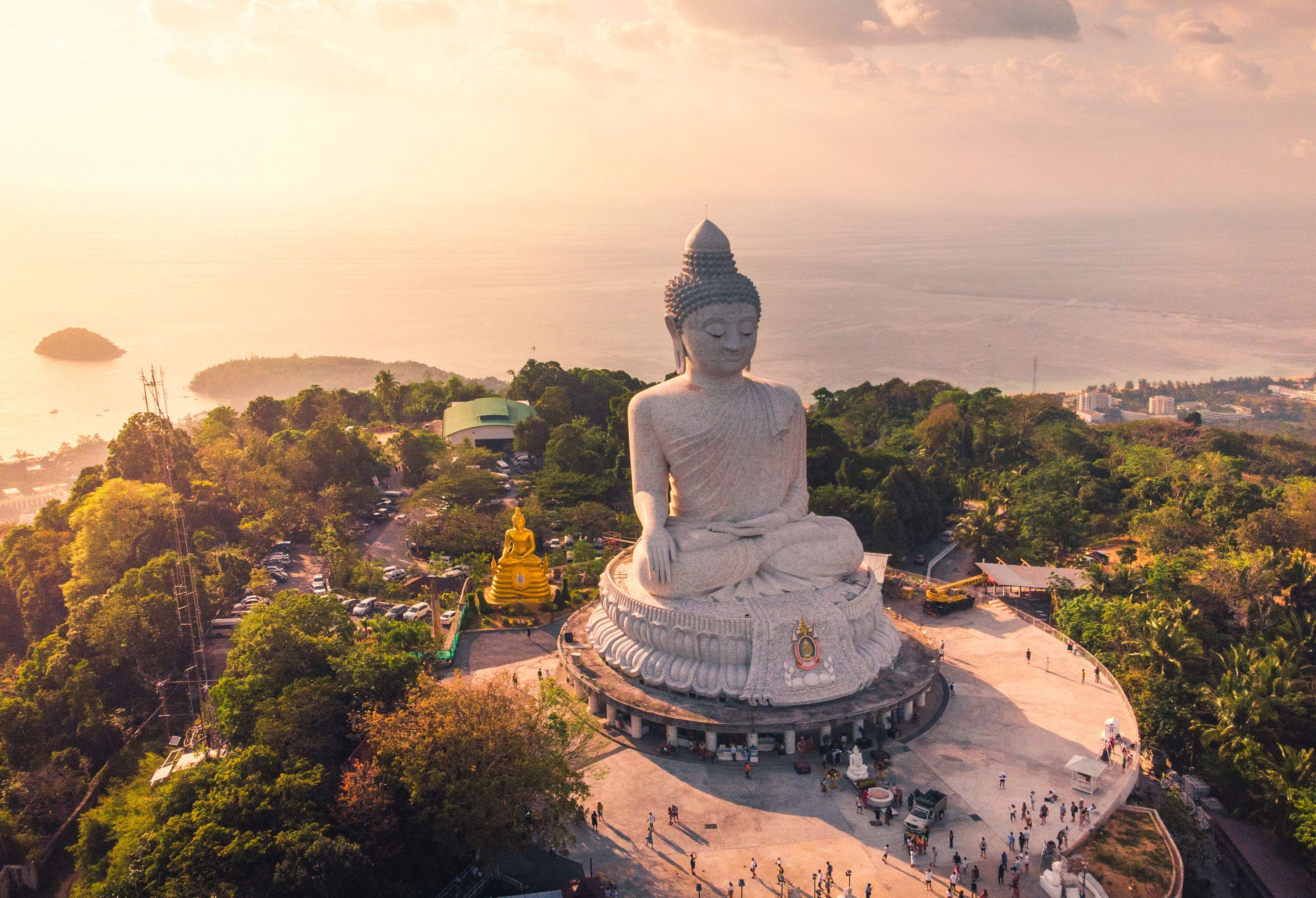 The Phuket Big Buddha statue sits at sunrise on a forested hill overlooking the sea.