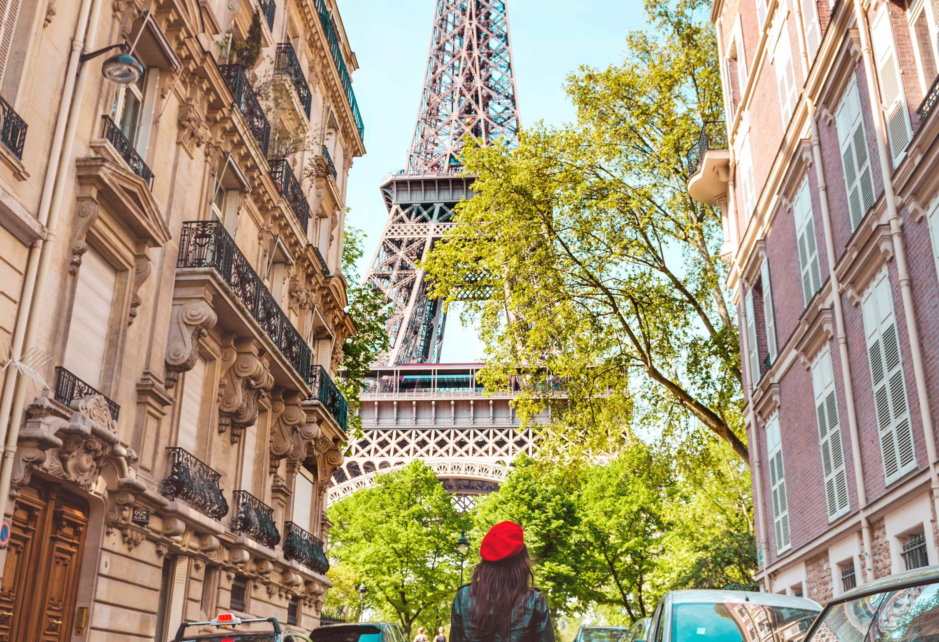 A woman in casual clothes walks on a street bordered by parked cars and adjacent buildings while staring at the Eiffel Tower.