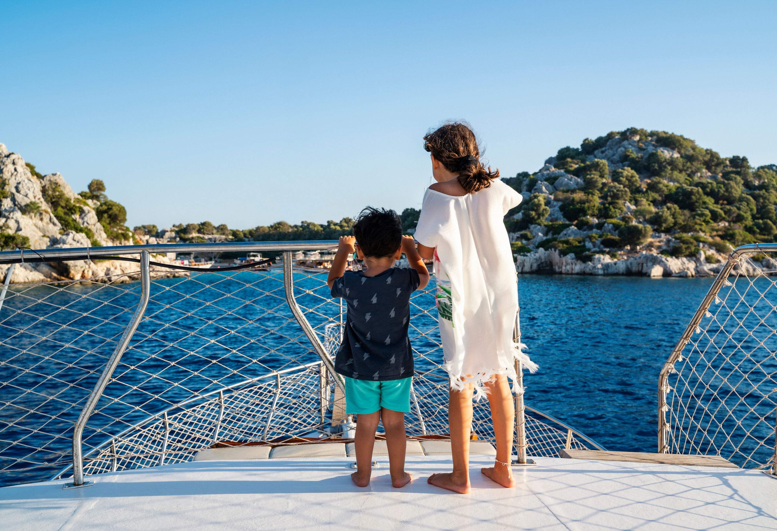 A little girl and her brother embark on an adventure on a sailboat, journeying towards an island with rugged slopes that loom in the distance.