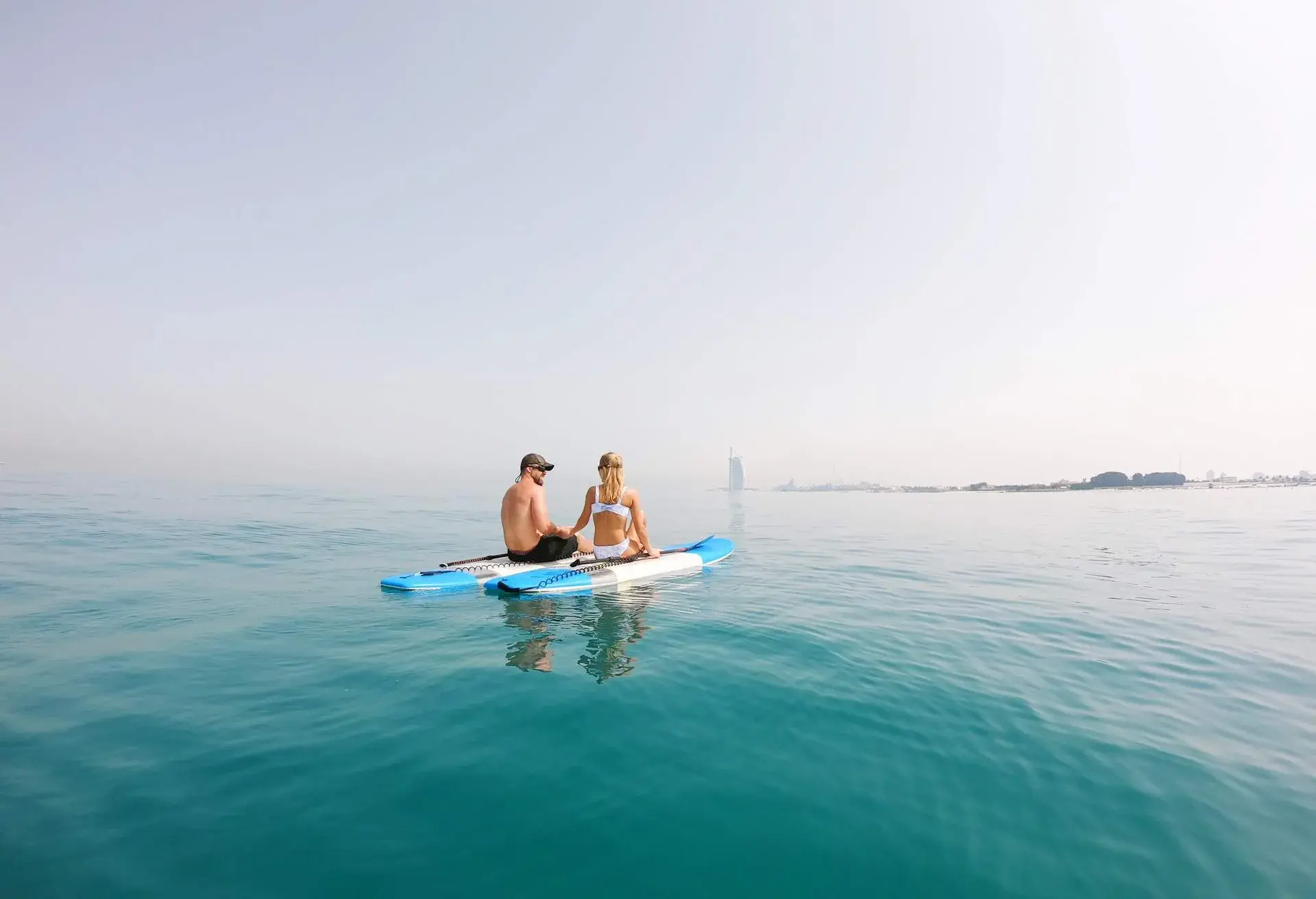 Couple sitting in the middle of the ocean on a stand up paddle near a city