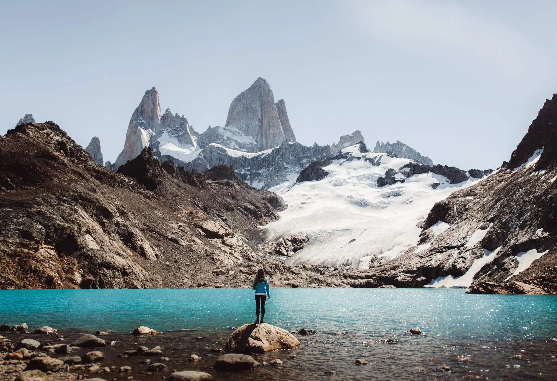 A person stands on a lakeside boulder gazing over the snow-covered valleys and mountain peaks.