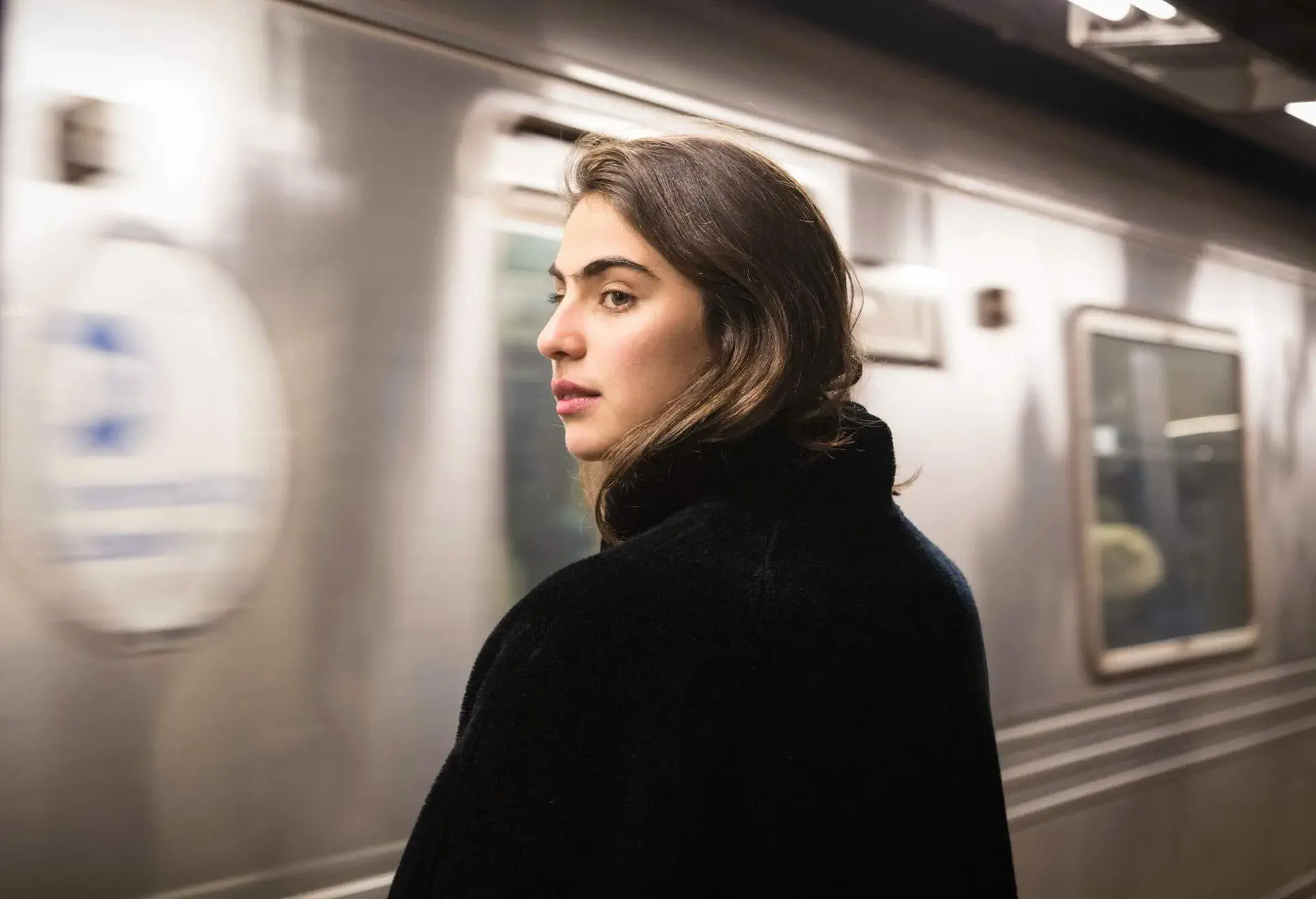 A brunette woman in a black jacket glances sideways against the backdrop of a moving train.
