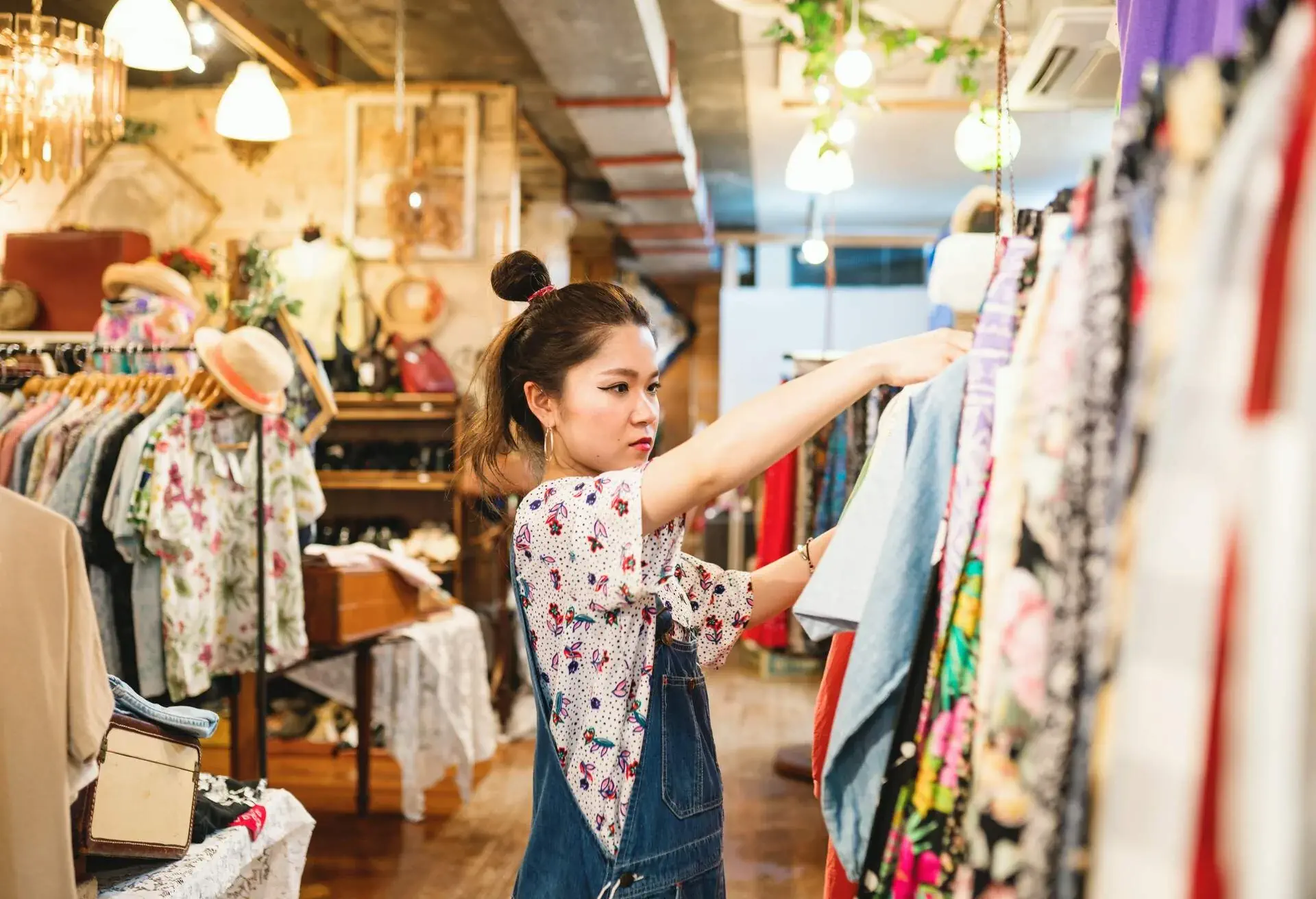 A woman in a floral shirt and denim jumper examines a dress in a clothing store.