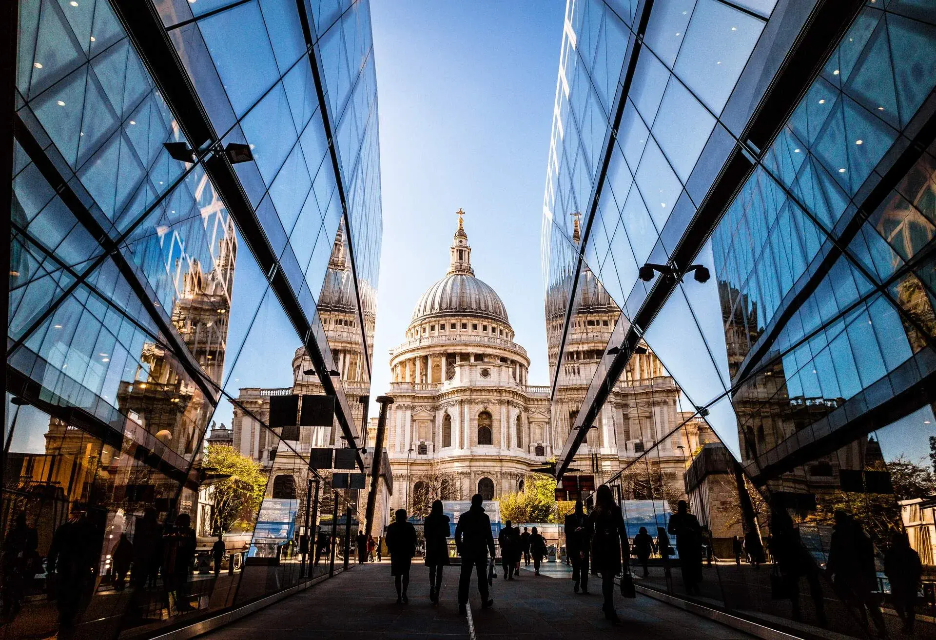People's silhouettes wandering across modern, futuristic structures of glass and steel that lead to a cathedral with a cross-topped dome.