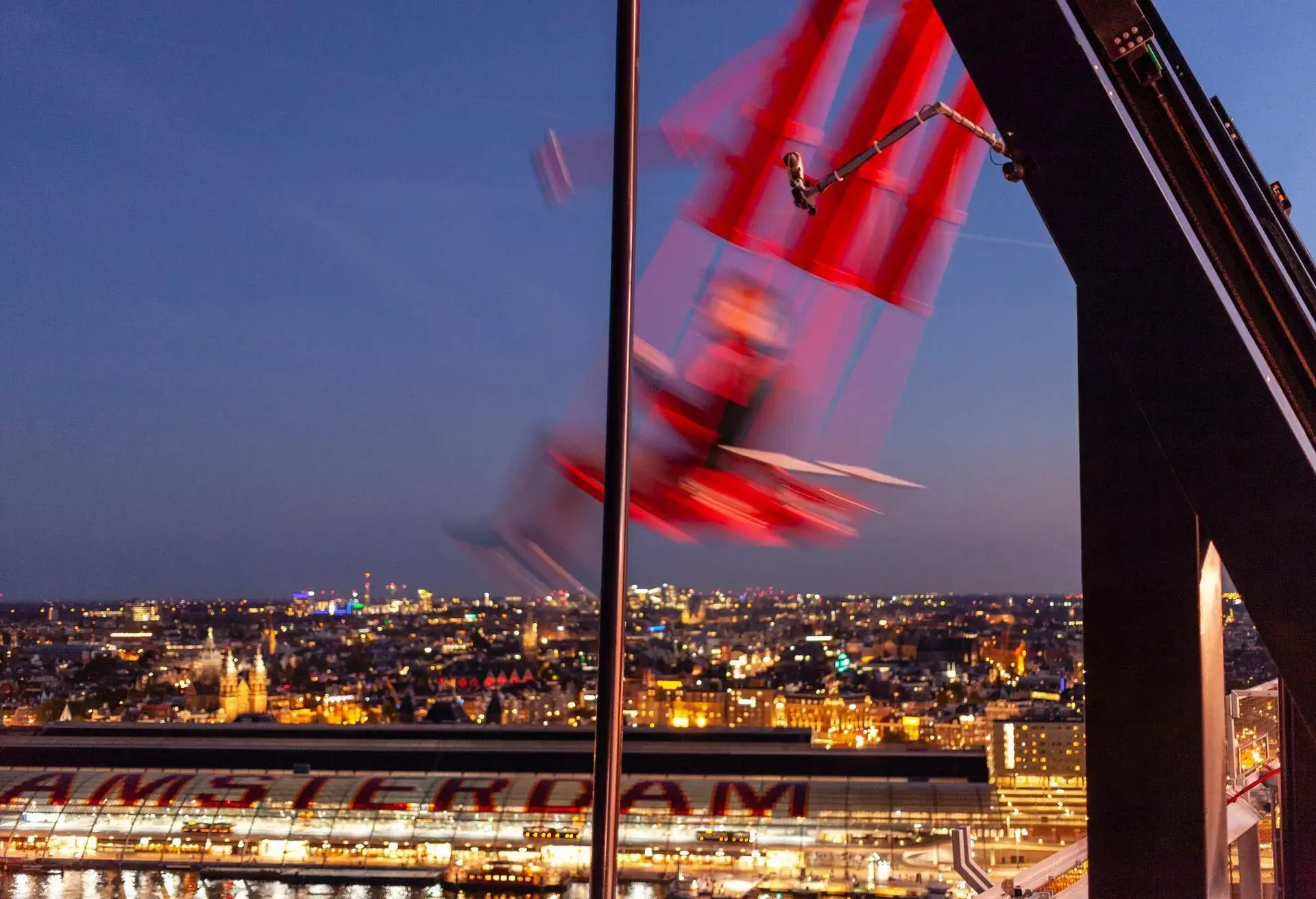 people sitting on a big red swing on the top of a building in Amsterdam at night with the skyline in the background