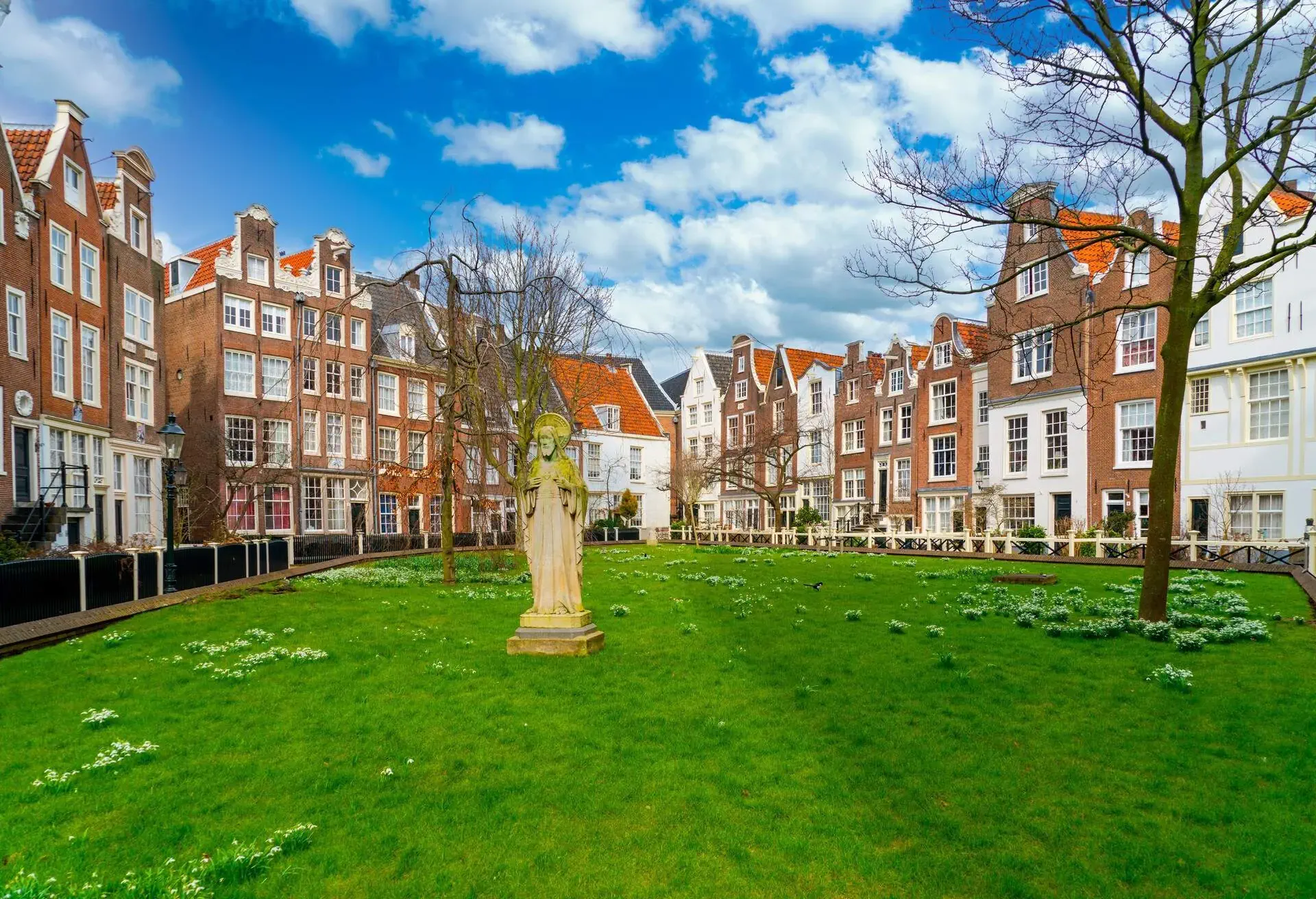 A statue in the middle of a park in Amsterdam surrounded by narrow buildings and with green grass and spring flower on the ground