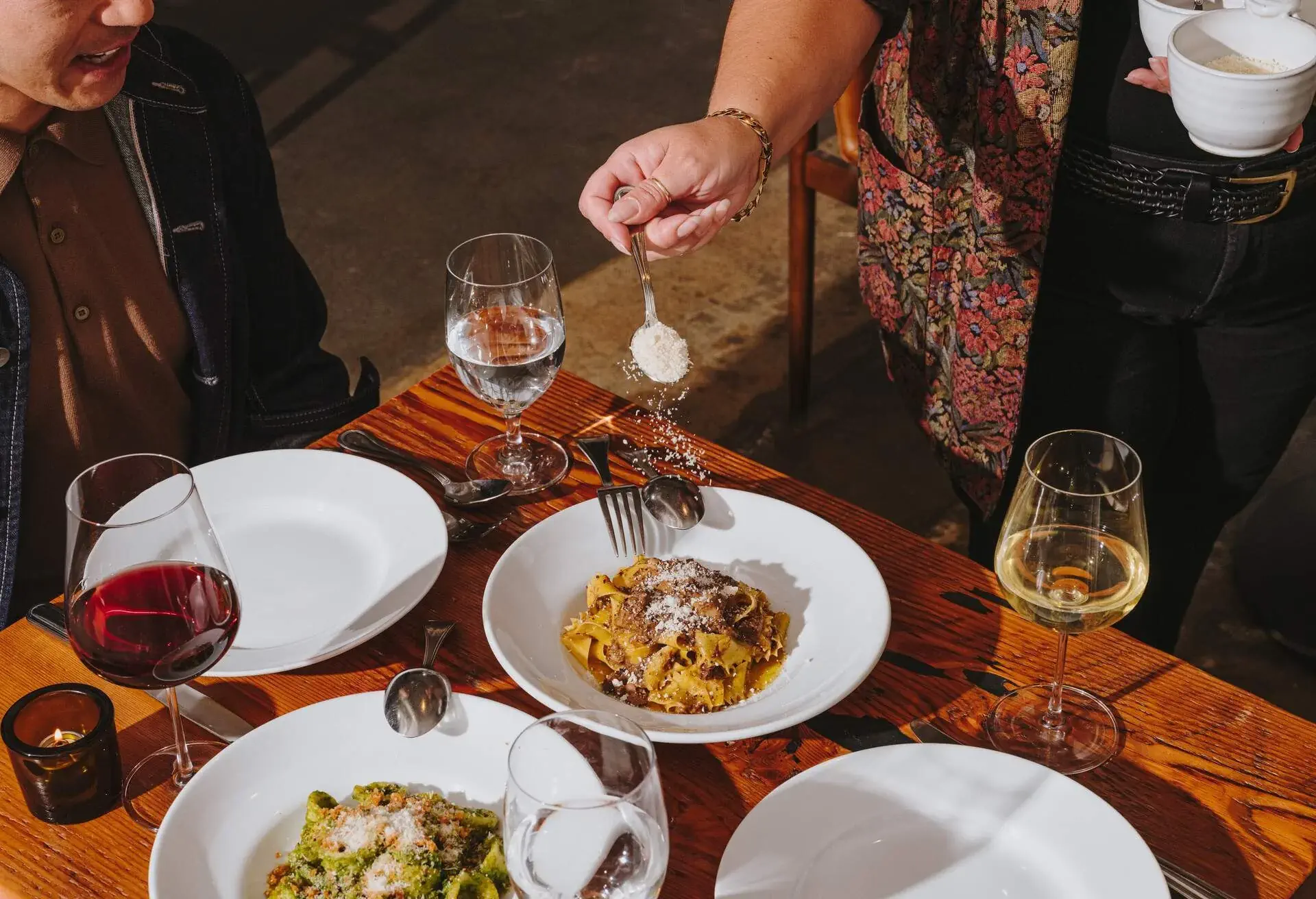 A woman dusting parmesan cheese over a plate of pasta at a customer's table.