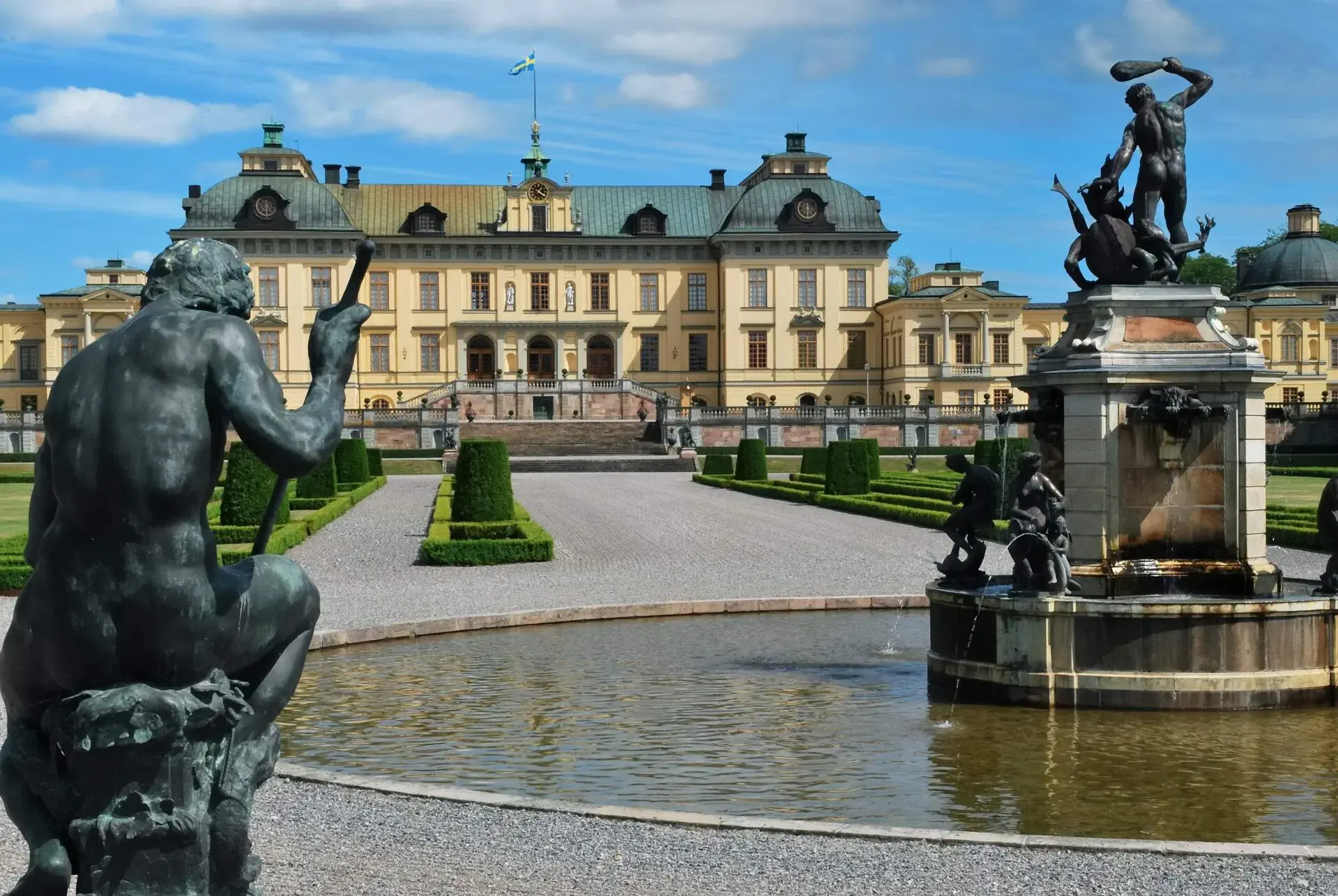 An elegant Baroque-style royal residence palace with a symmetrical façade, decorative sculptures, intricate details and a lush garden of shrubs with a fountain in the centre.