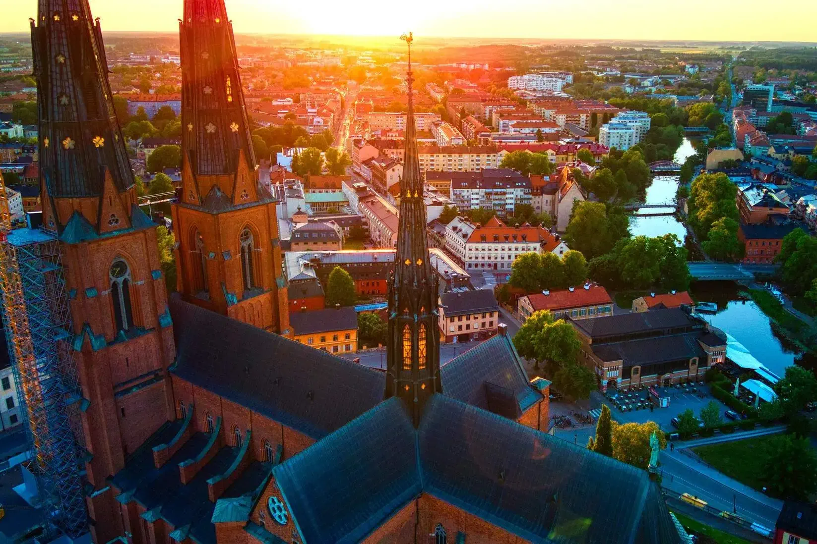 A sunlit cityscape from a church spire overlooks a river between the compact clusters of tall buildings.