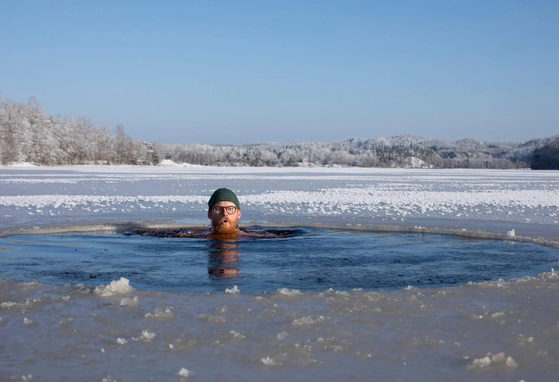 Bearded ginger man wearing hat and glasses swimming into ice cold water in a ice hole during a winter swimming session