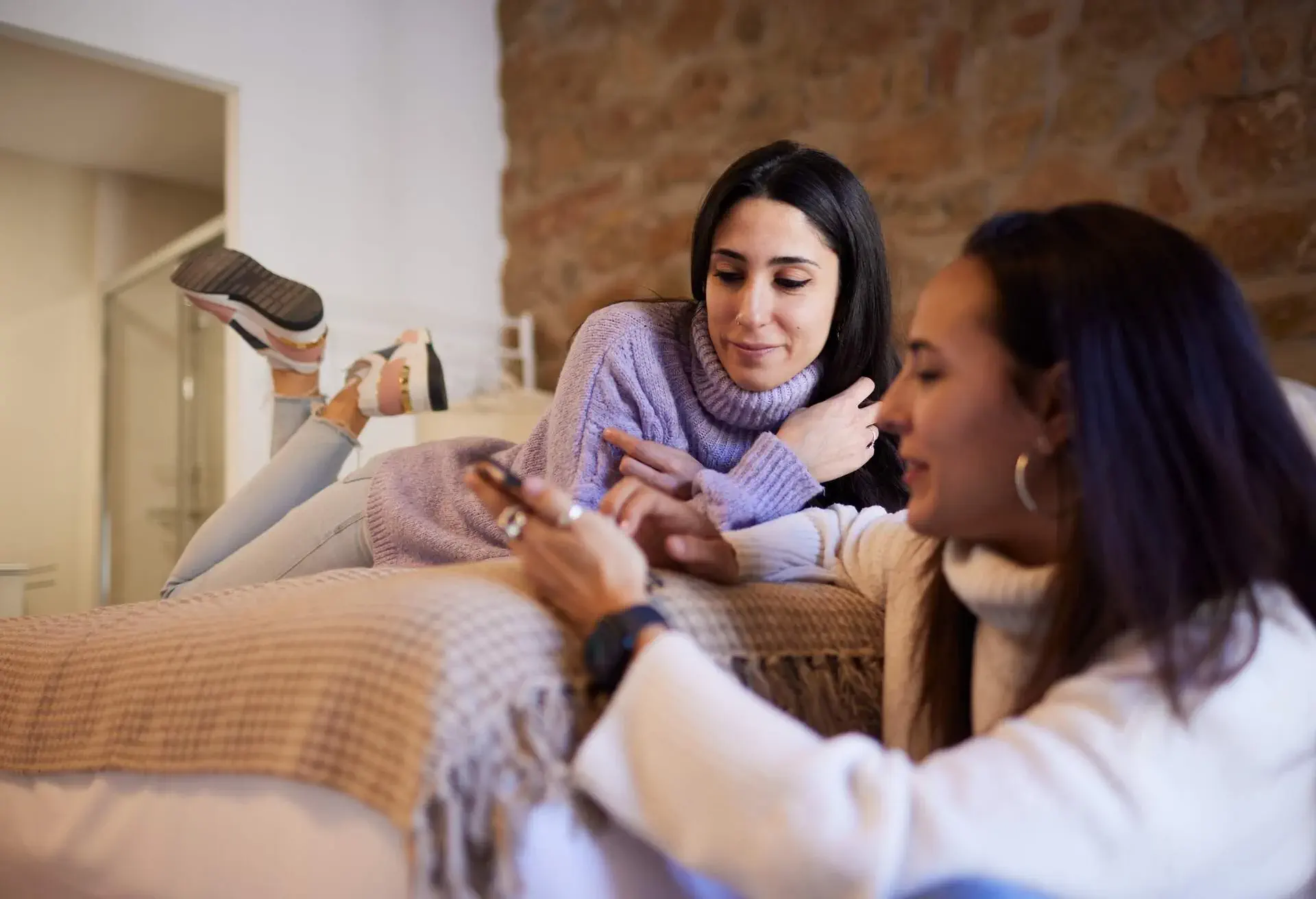 One friend lies on the bed facing down while the other sits on the floor and shows her a smartphone screen, both fully engaged in the content they are viewing together.