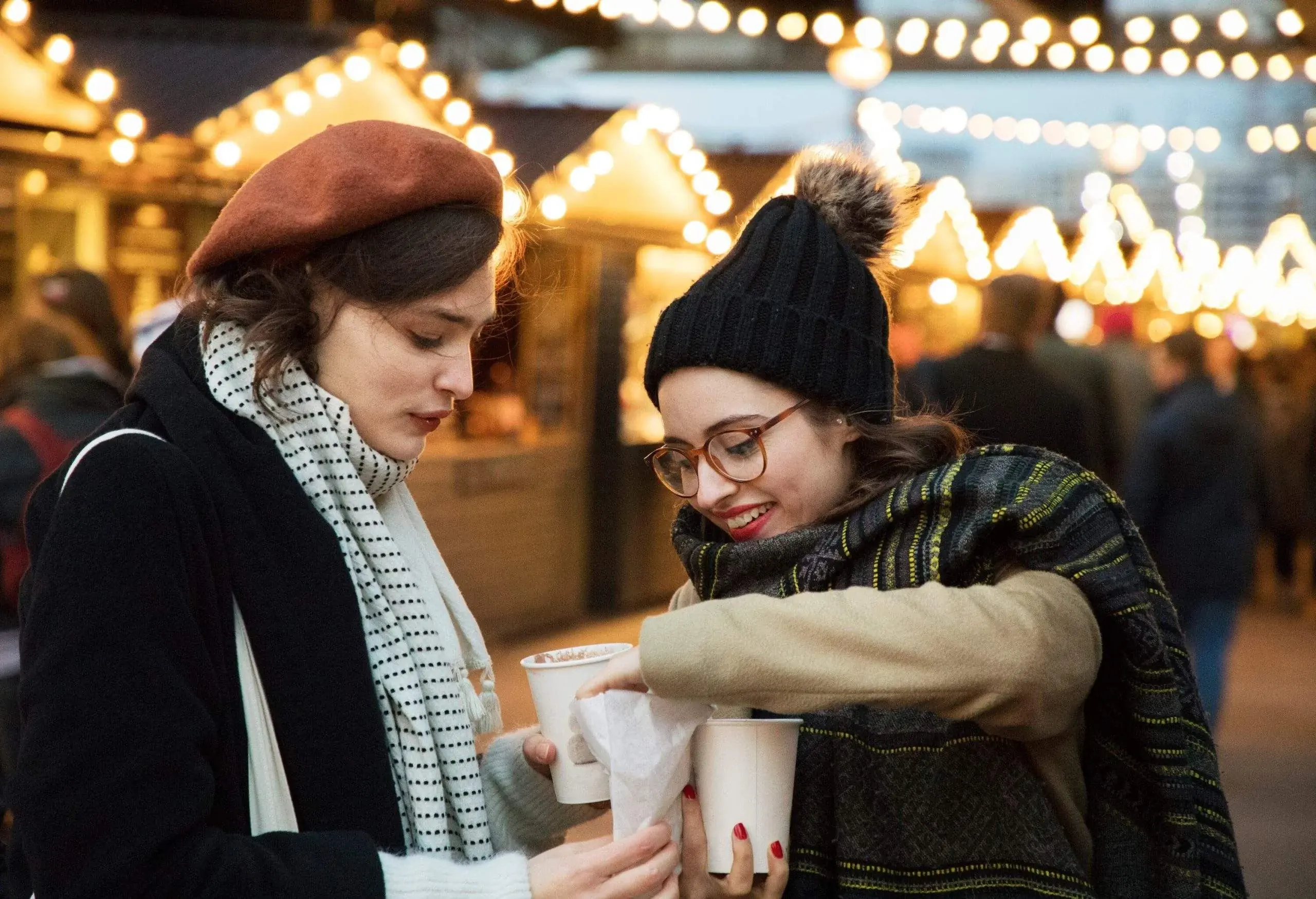 Two women sharing treats and drinking hot chocolate at Christmas market, wearing wooly hats and scarves