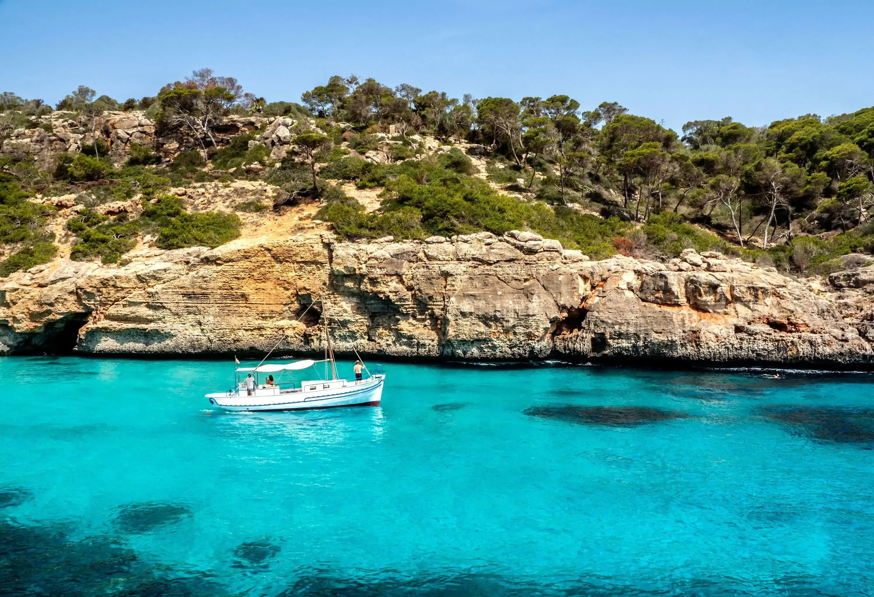 A boat cruising in a blue sea bordered by a rock cliff topped with trees.