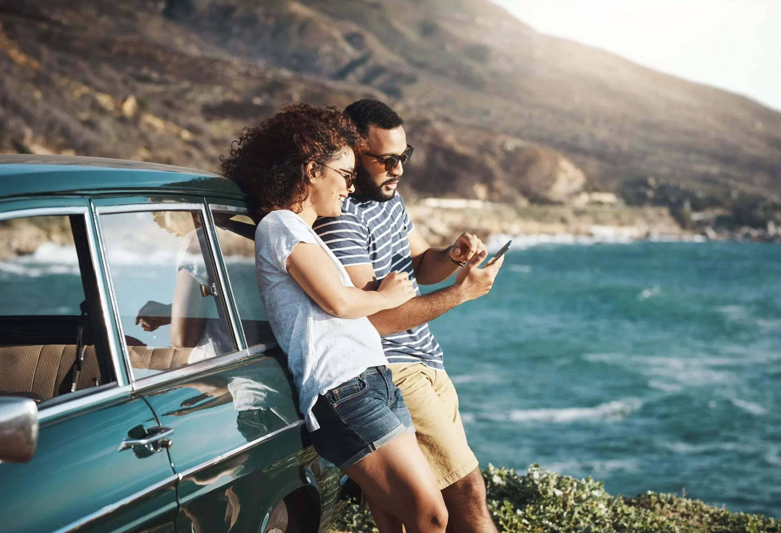 A woman and a man leaning on a blue car, looking at a smartphone, next to the sea
