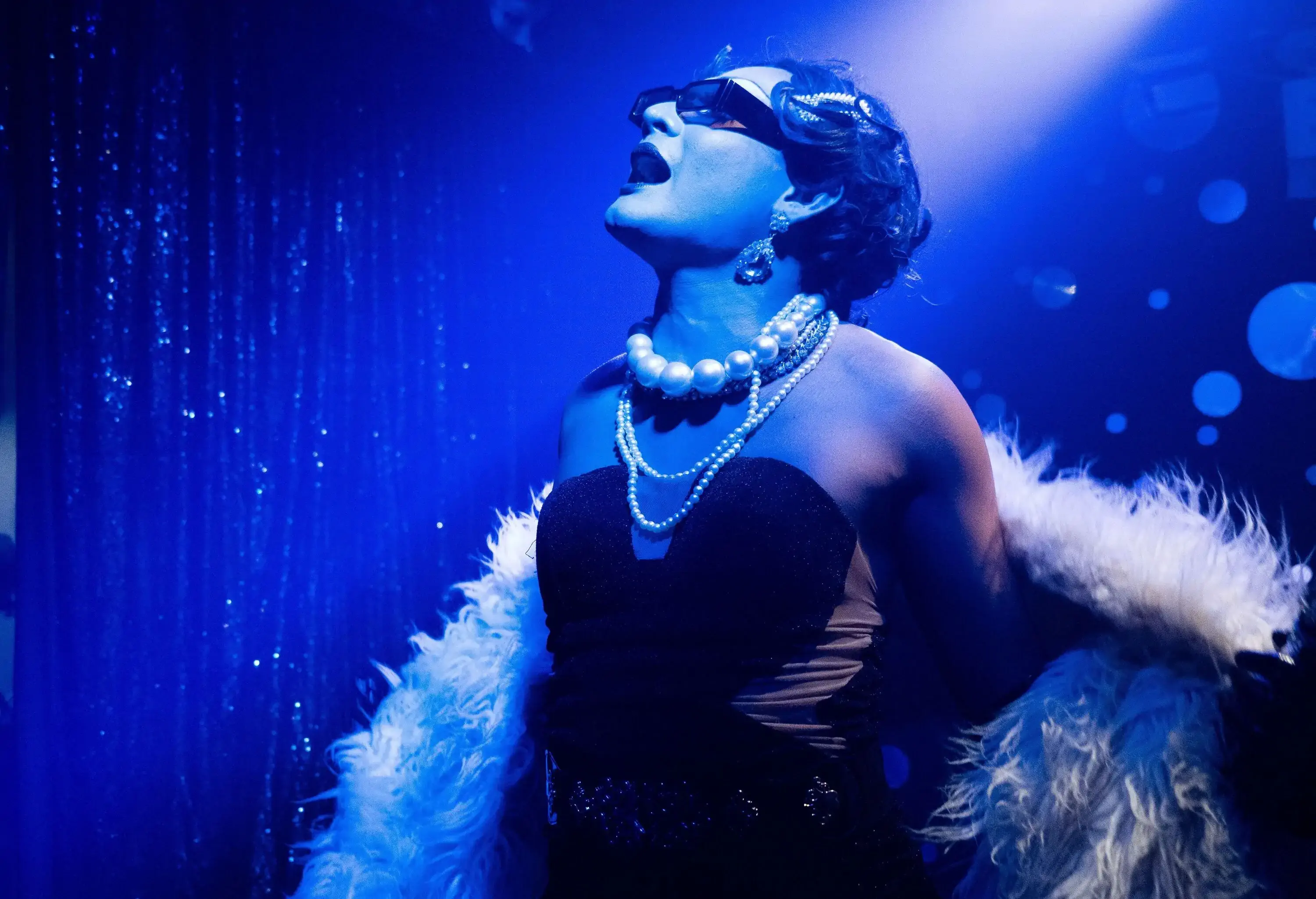 A drag performer in black dress, feathered cape, and pearl necklaces sings on a stage with blue lights.
