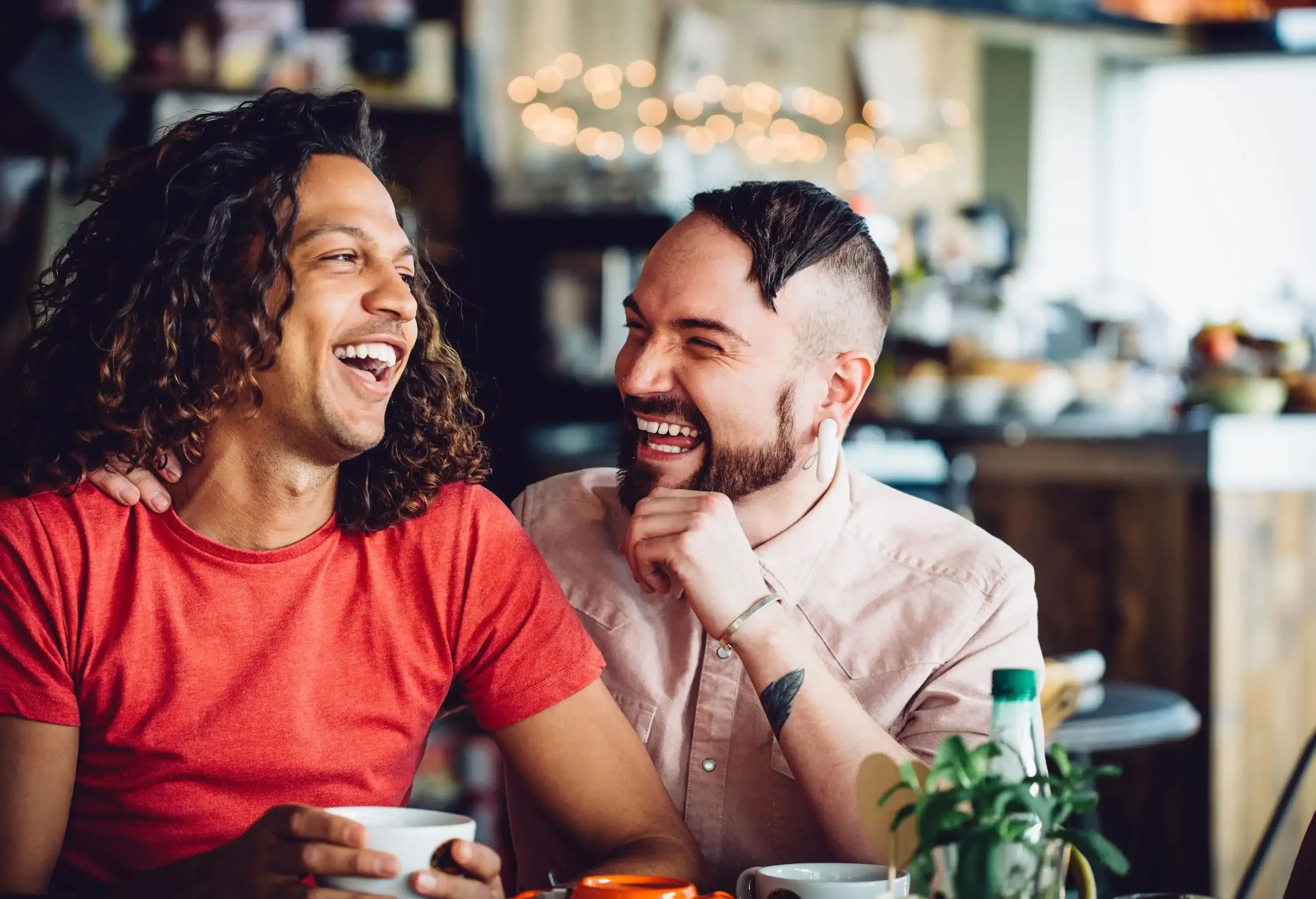 A gay couple cosy up in a coffee shop, they are laughing together and enjoying each others company while drinking coffee in a modern/trendy cafe.