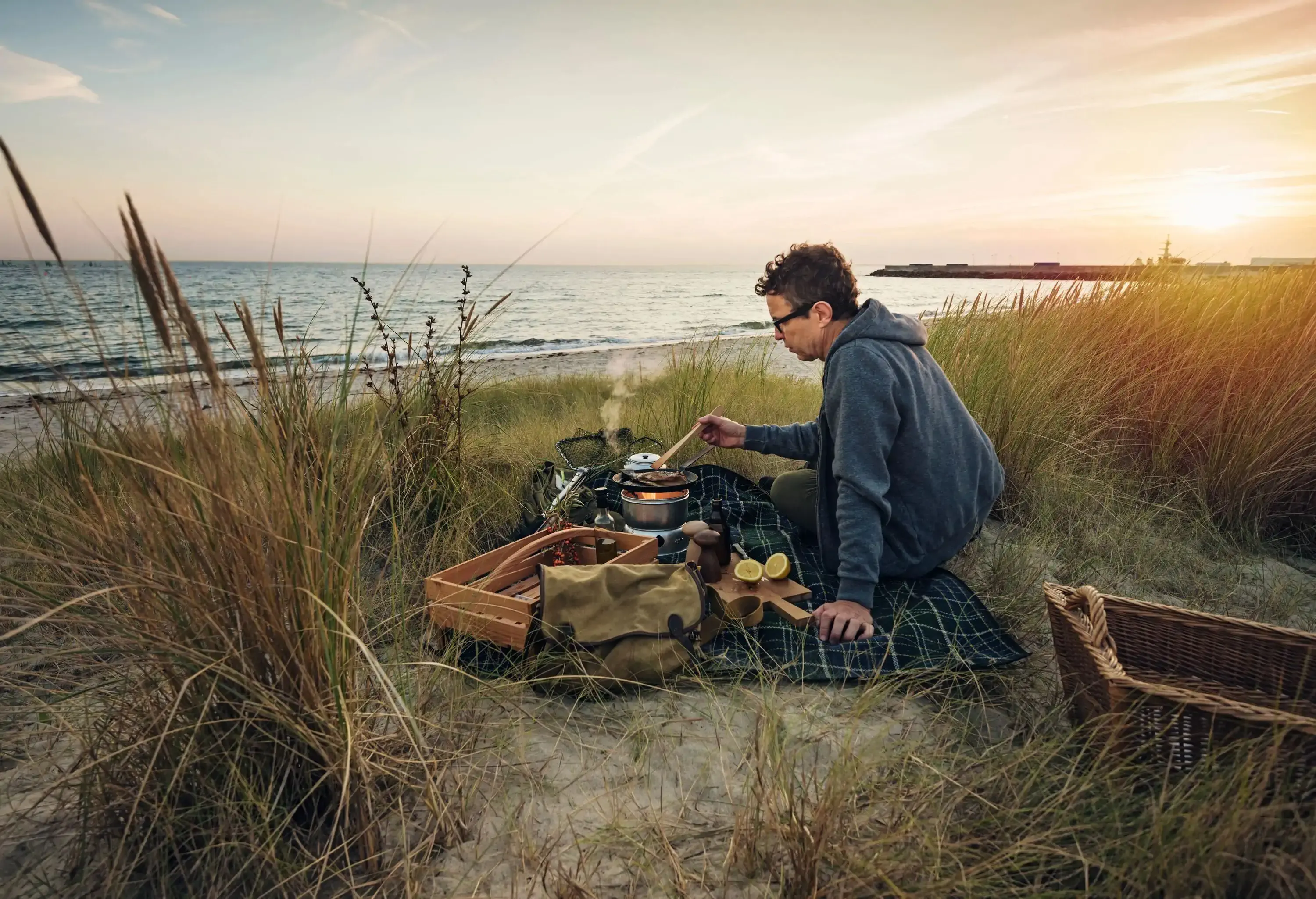 A lone man camping on the grassy shore of the beach, cooking his dinner before twilight.
