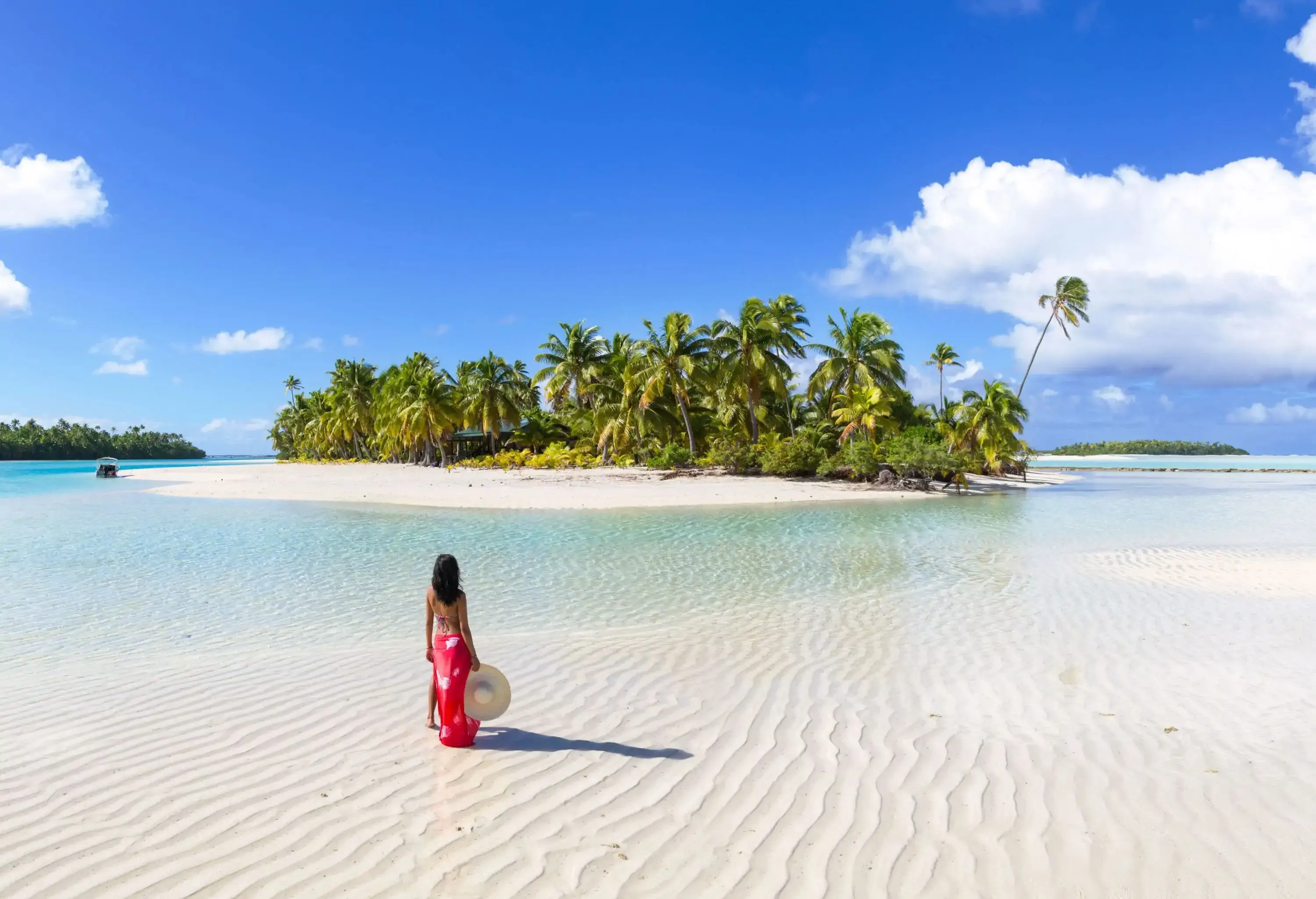 A woman wearing a sarong standing on a pristine beach holding her hat.