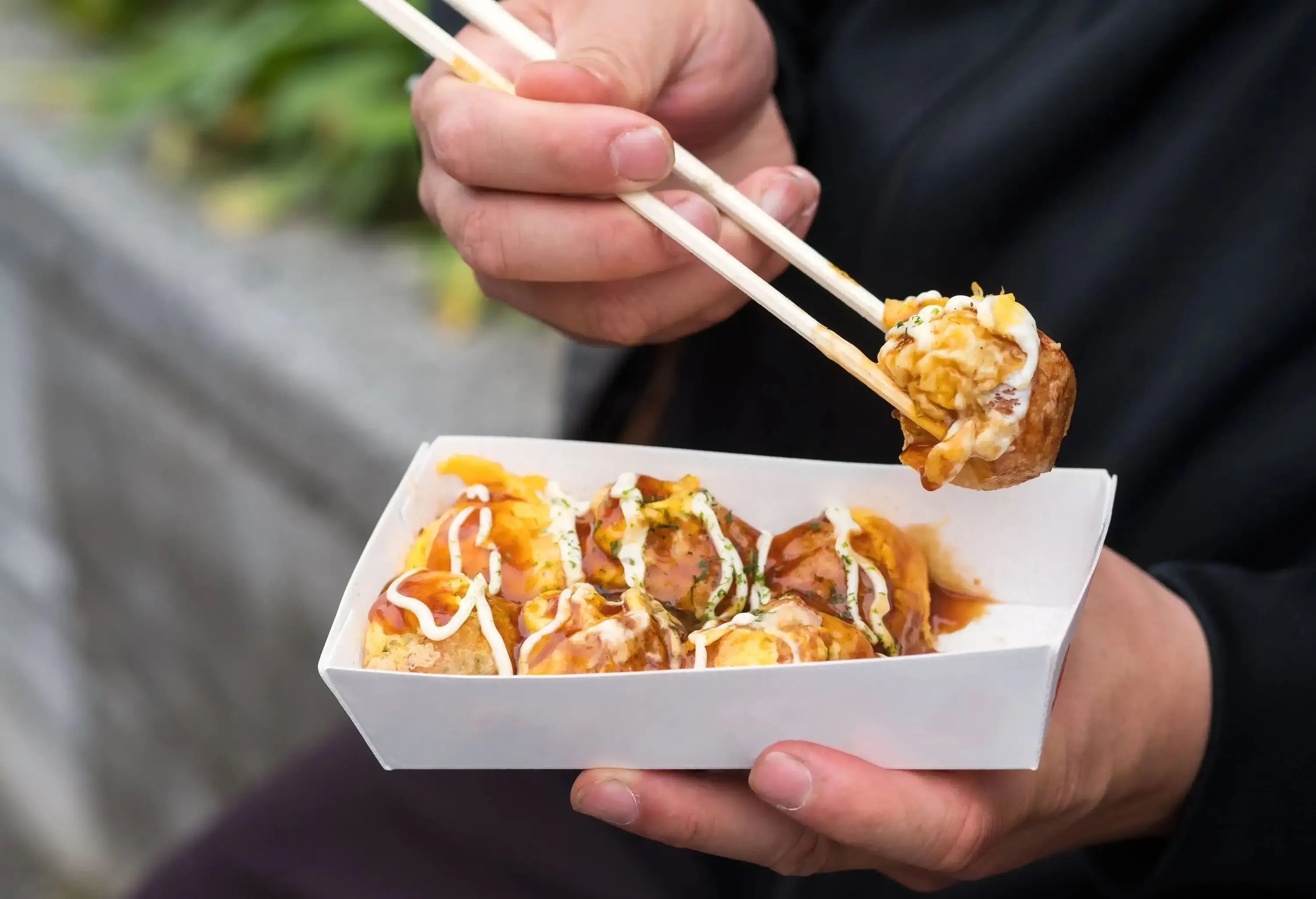 A man picking up a takoyaki ball from a white carton box with a pair of chopsticks.