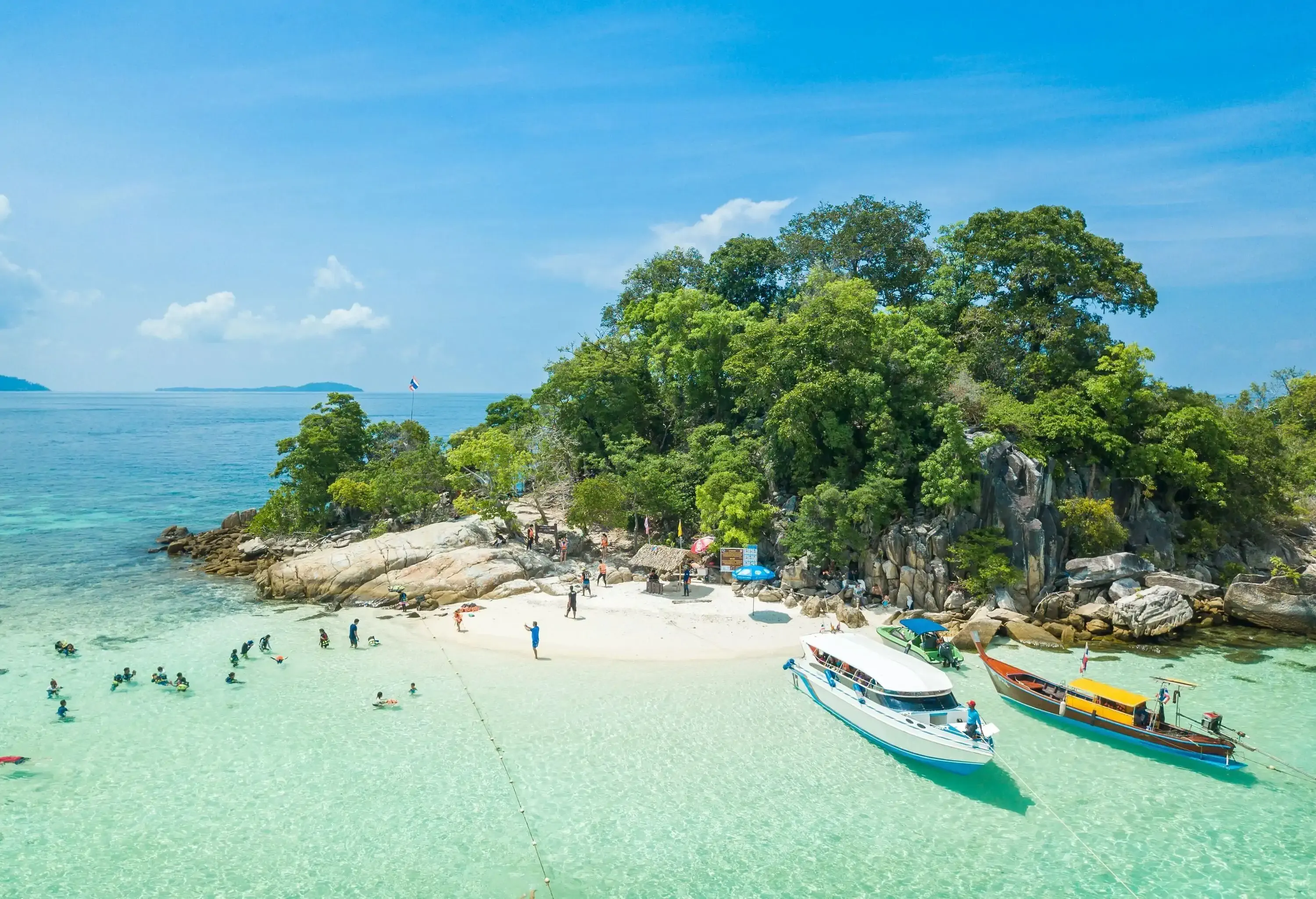 Aerial view of a paradise with a white sand beach and rugged island surrounded in tall green trees by the turquoise clear water sea.