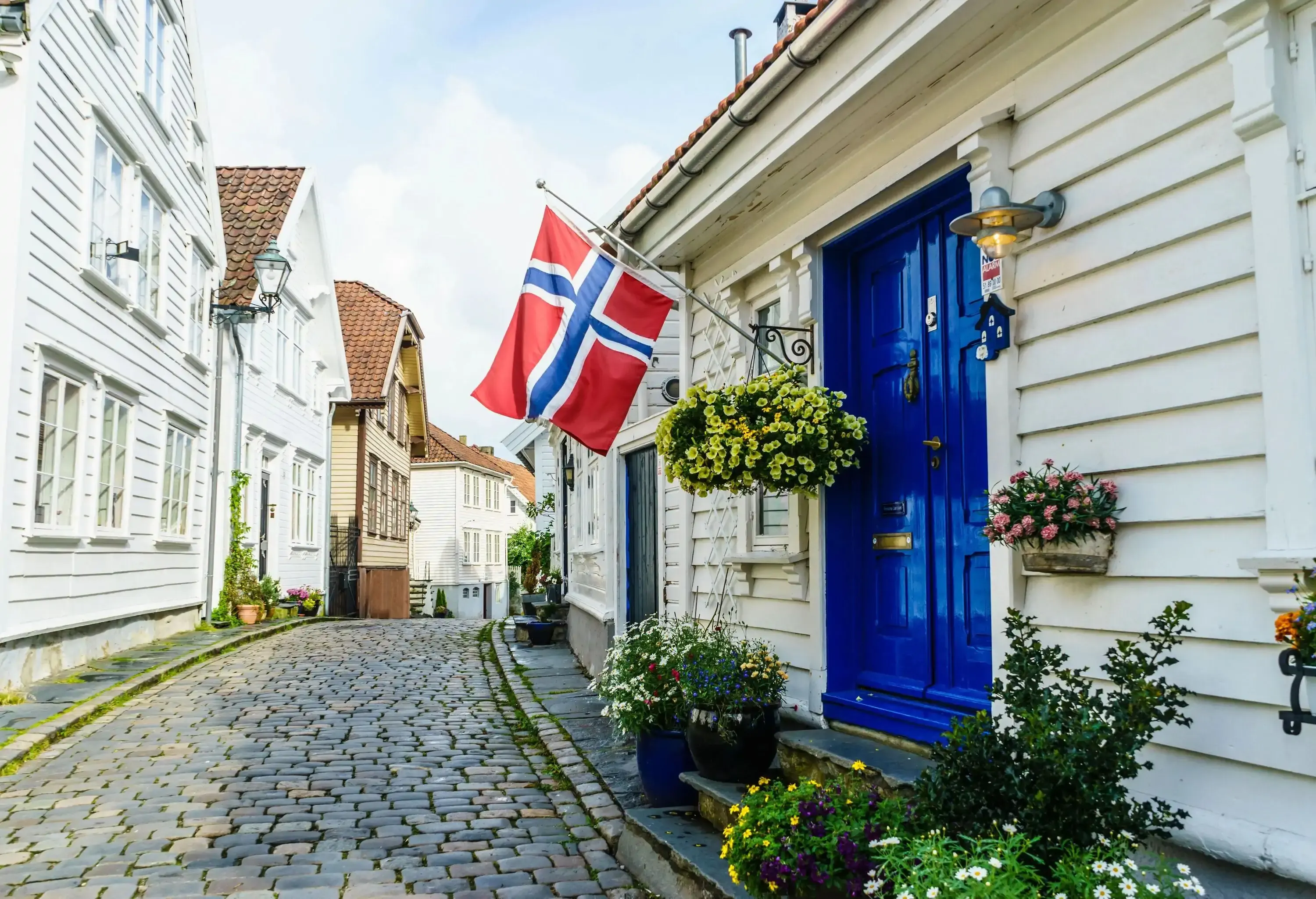 the Norwegian flag outside of an idyllic wooden house with a blue door on a small cobble alley