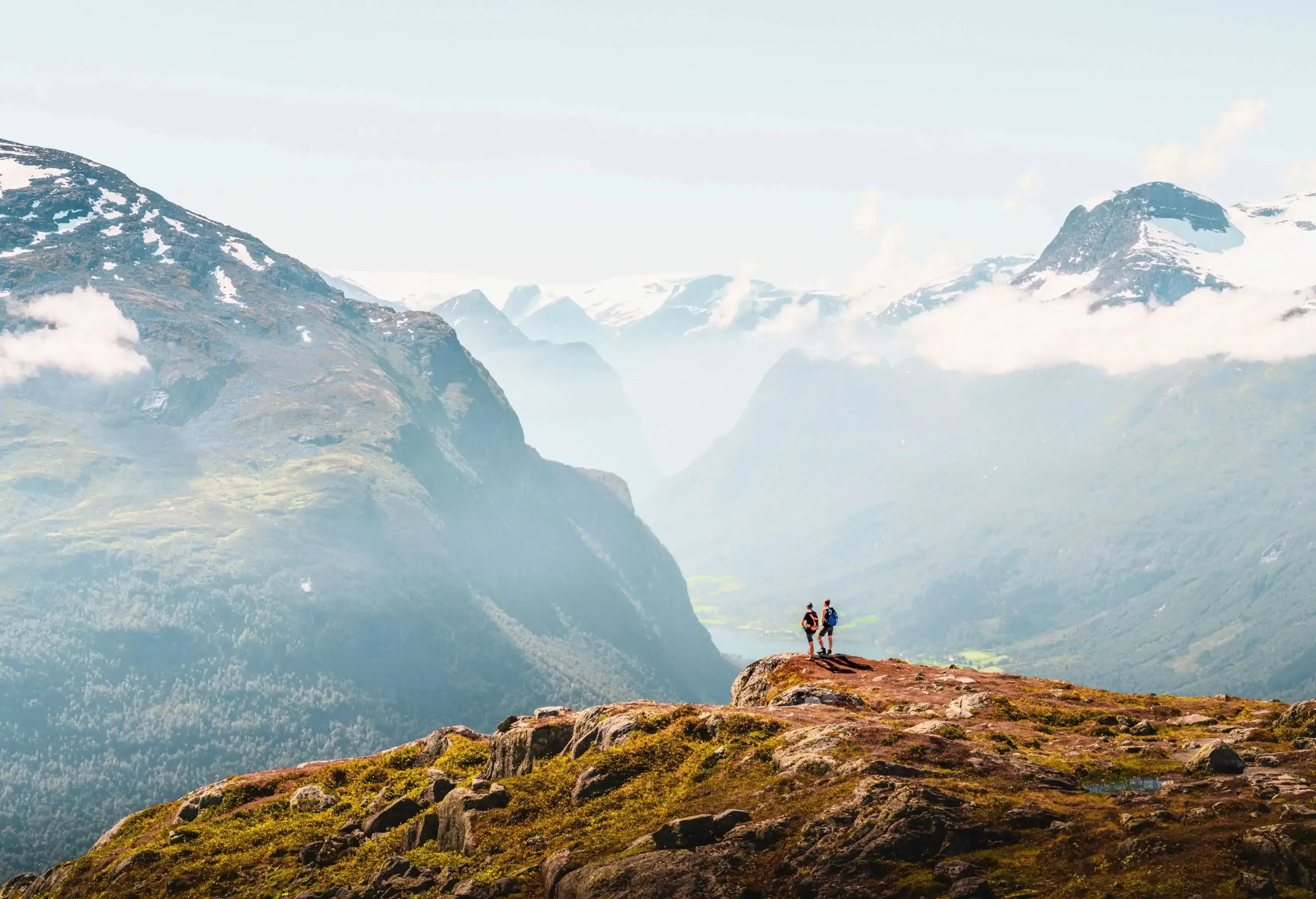 Two hikers stand on the cliff's edge, viewing the snow-capped mountain peaks against the sky in the distance.