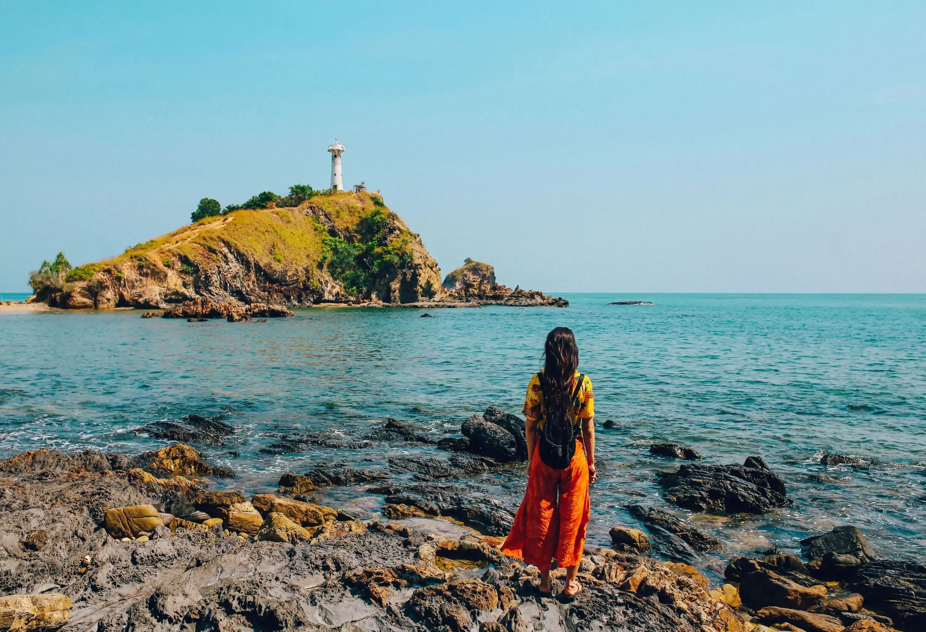A woman standing on a rocky shore, staring at a lighthouse perched on an island.