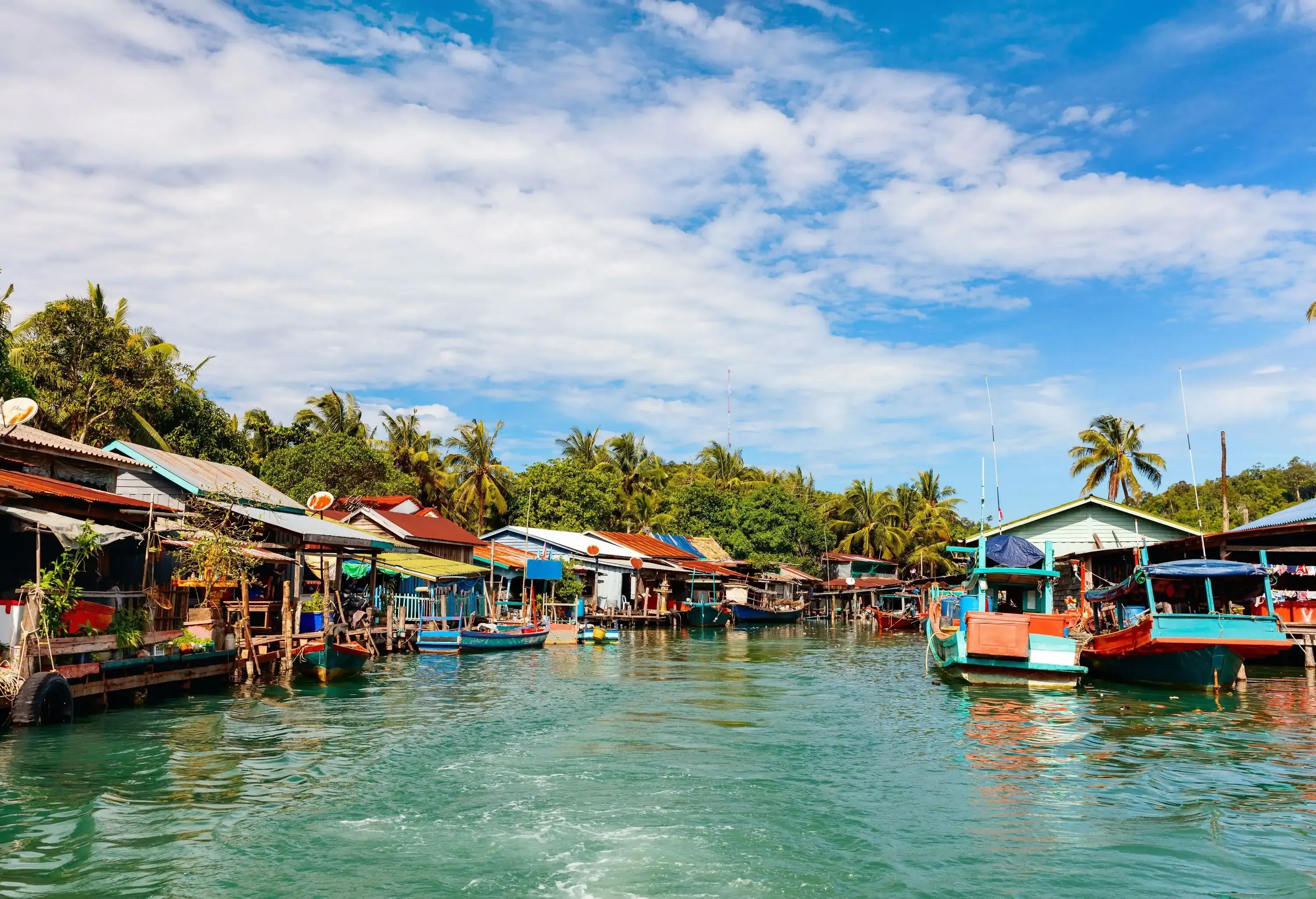 A traditional floating village on Koh Rong Island, featuring stilted houses with boats moored beside them, all backed by a lush array of tropical trees.
