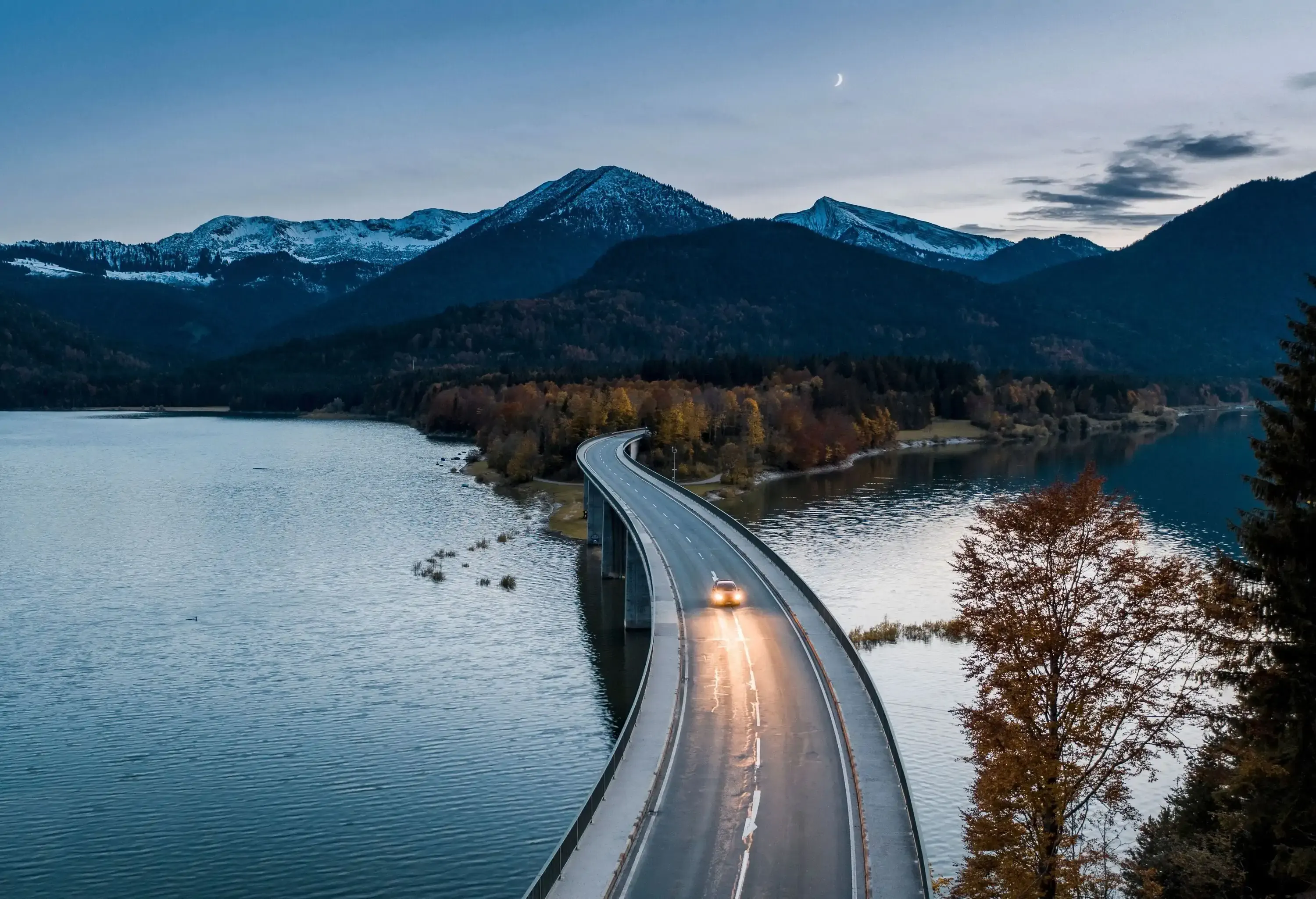 A car with its front light on drives along a curvy bridge across a broad river surrounded by steep snow-capped mountains.