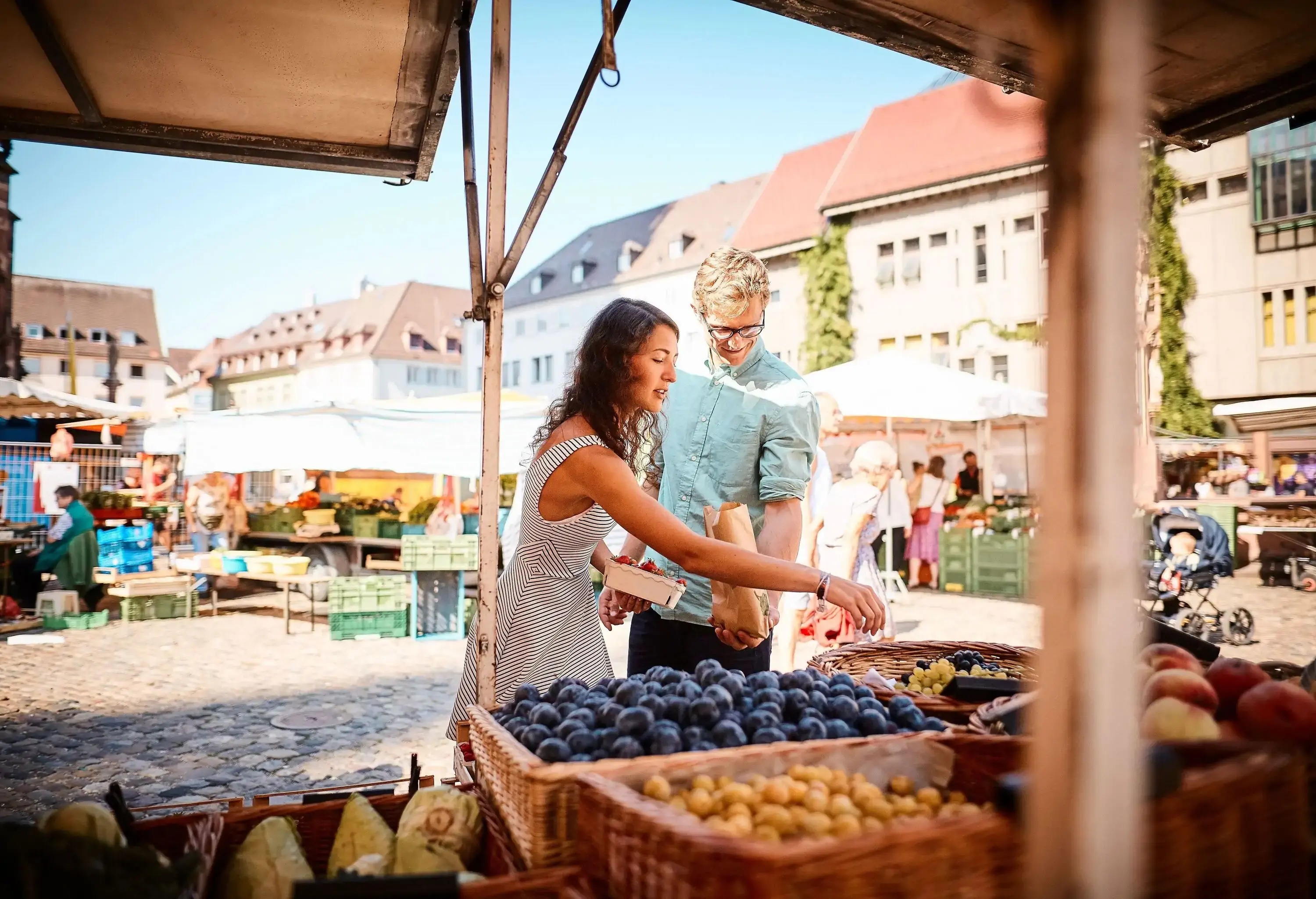 A couple picks some grapes in a fruit stall on the market surrounded by buildings.