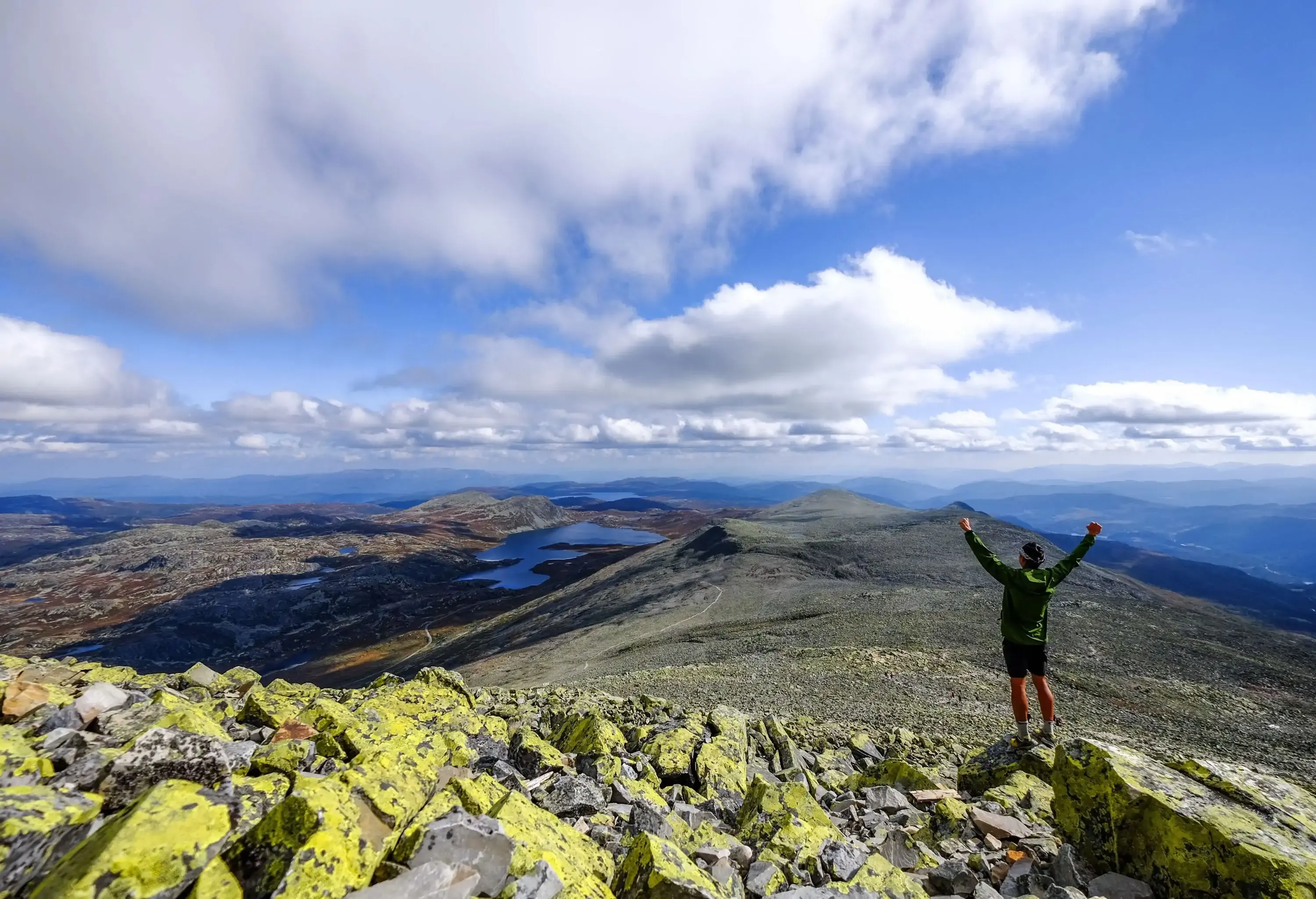 A hiker standing on a rock with arms raised looking over the vast mountainous area.