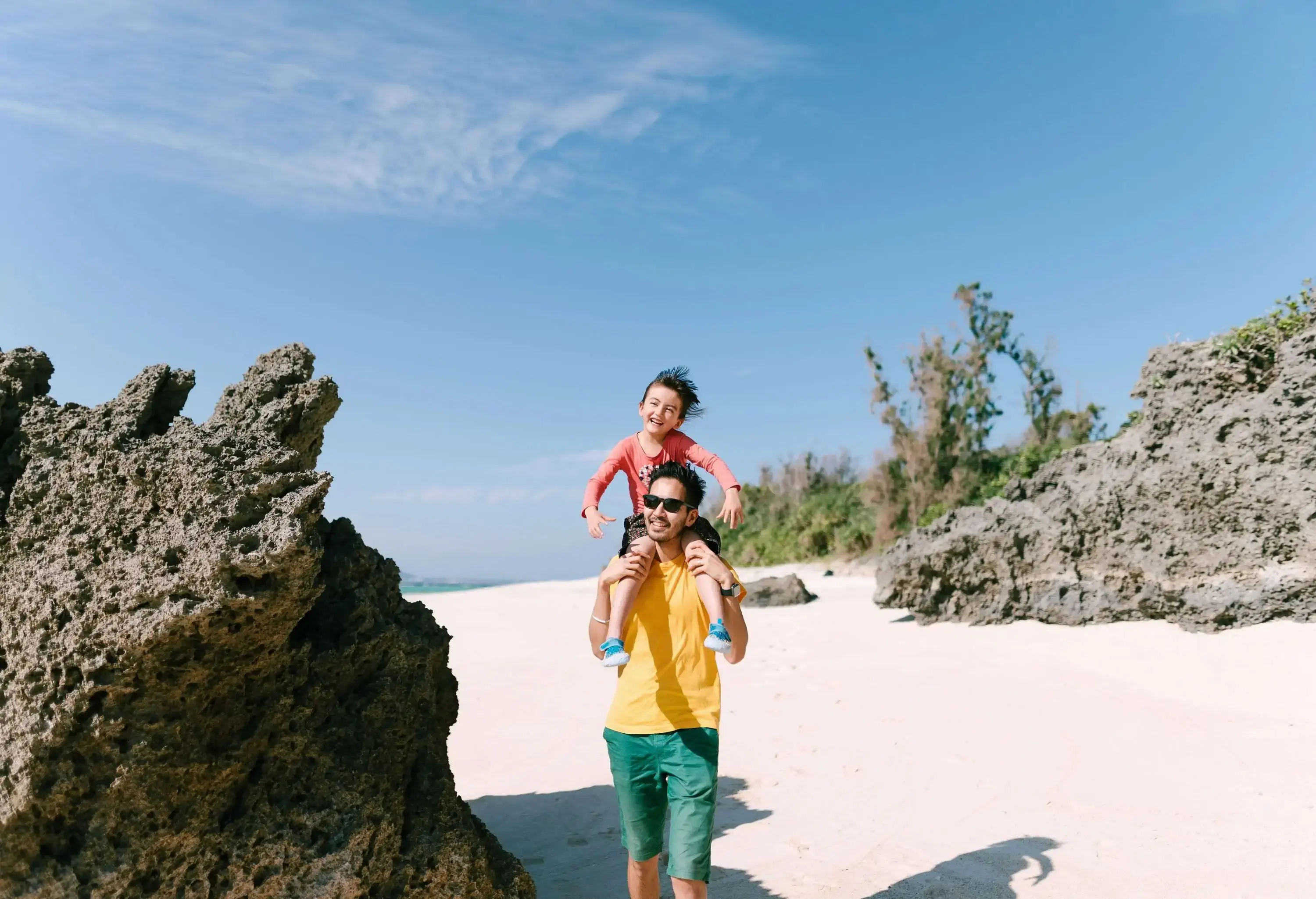 A father carrying his son on his shoulder as he walks the white sand beach during the day.