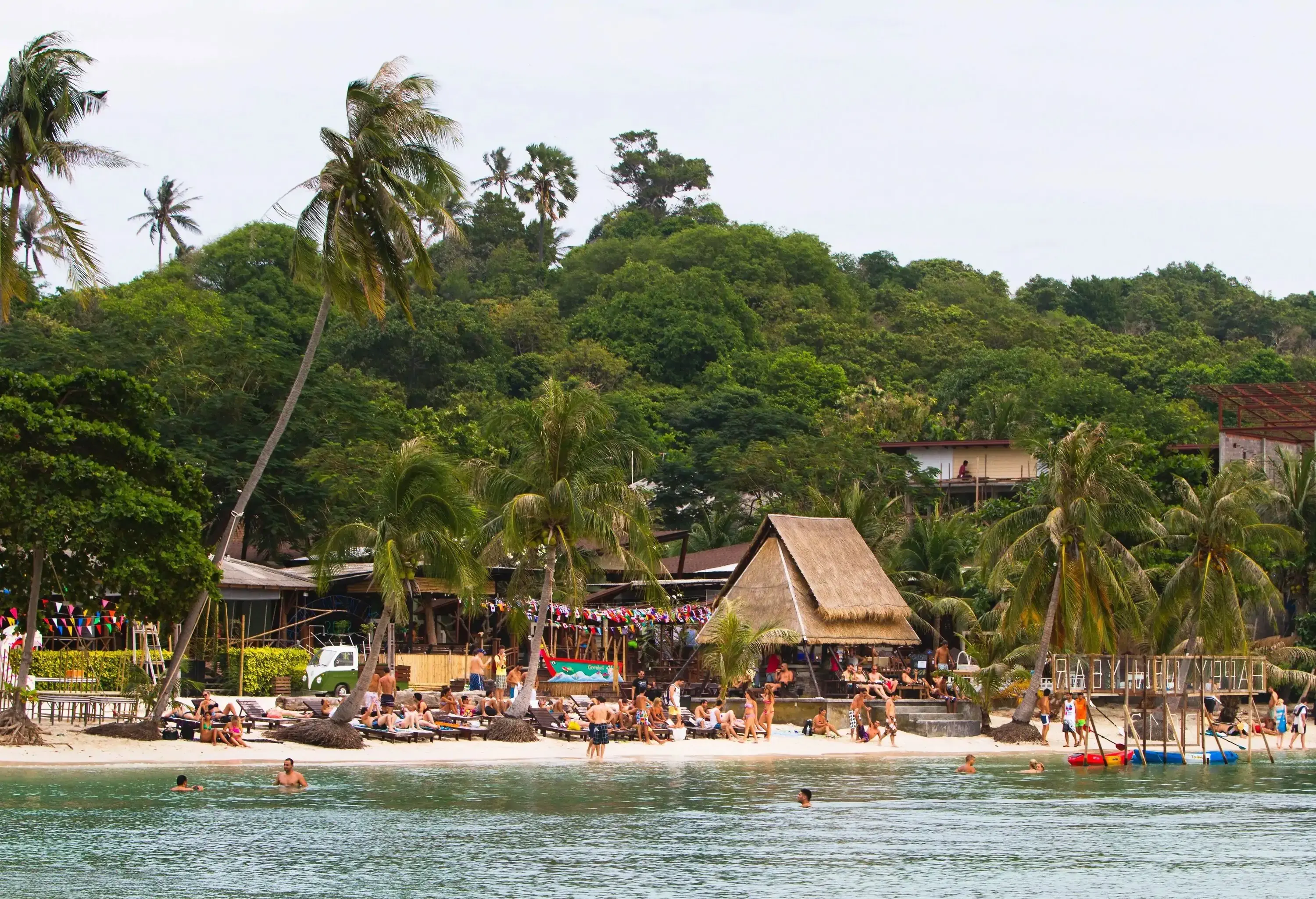 People on white sand shore by the sea lined with resort houses surrounded by tall green trees.