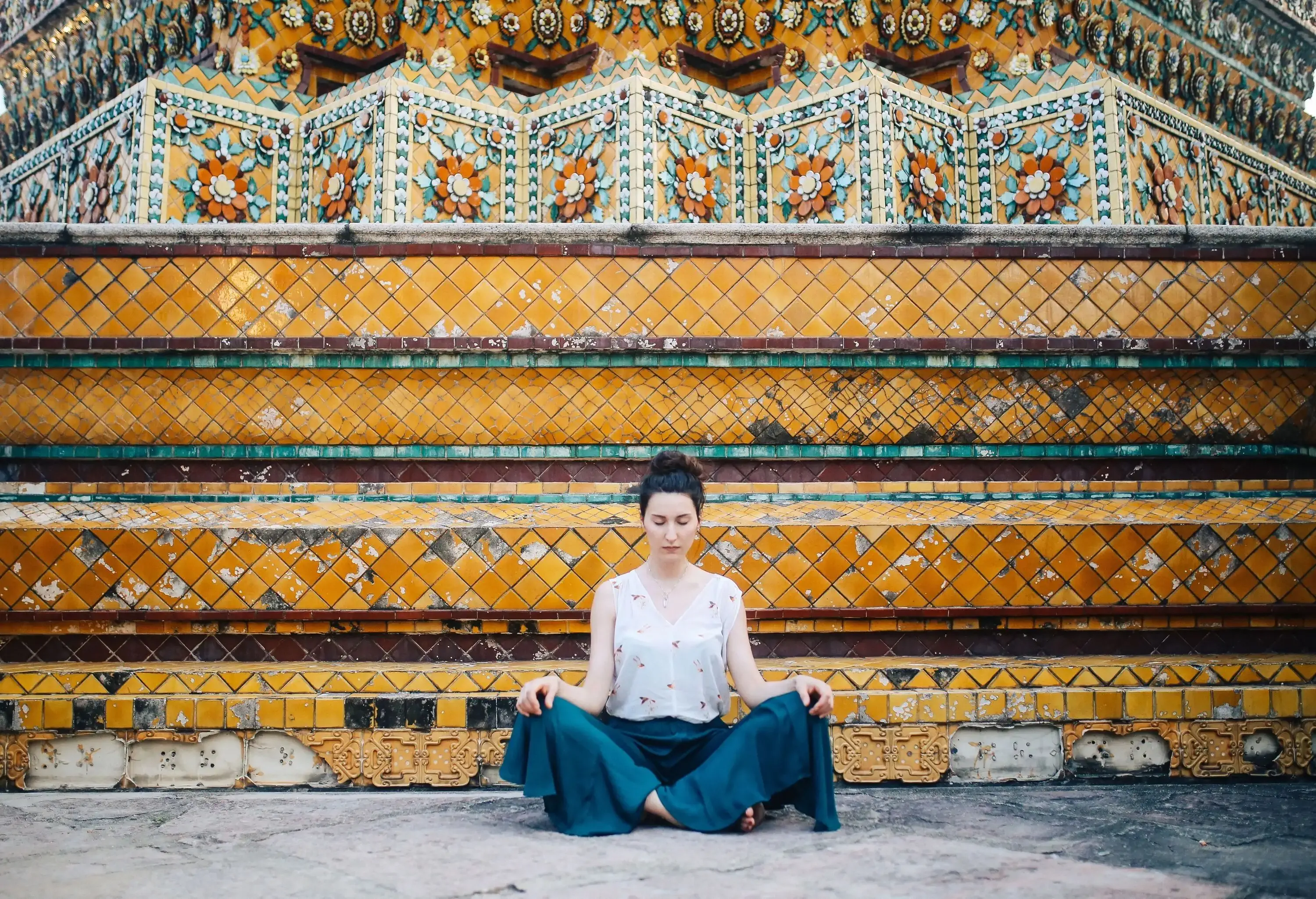 A woman sits and meditates against a tiered wall adorned with mosaics of colourful tiles.