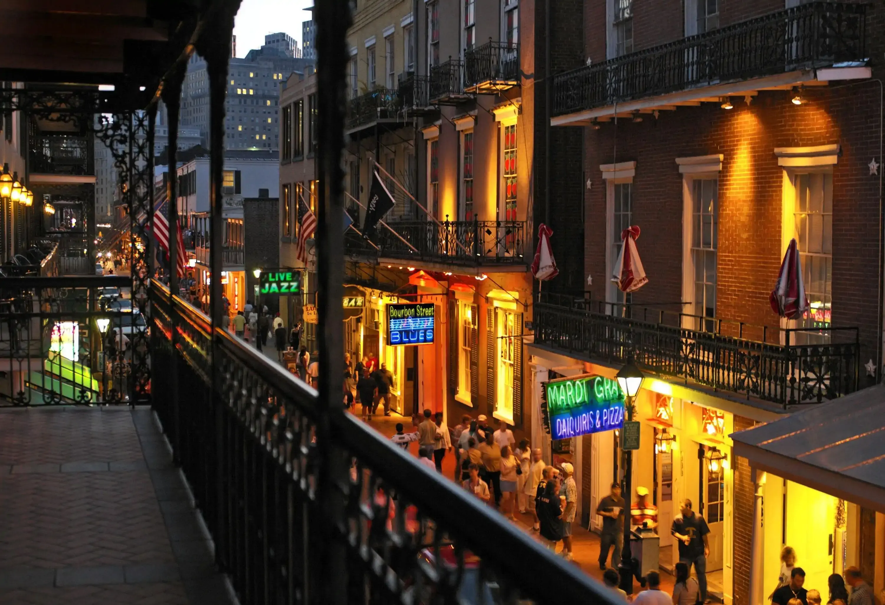 An apartment balcony overlooks a crowded sidewalk along adjacent apartment buildings with illuminated commercial establishments on the ground floor.
