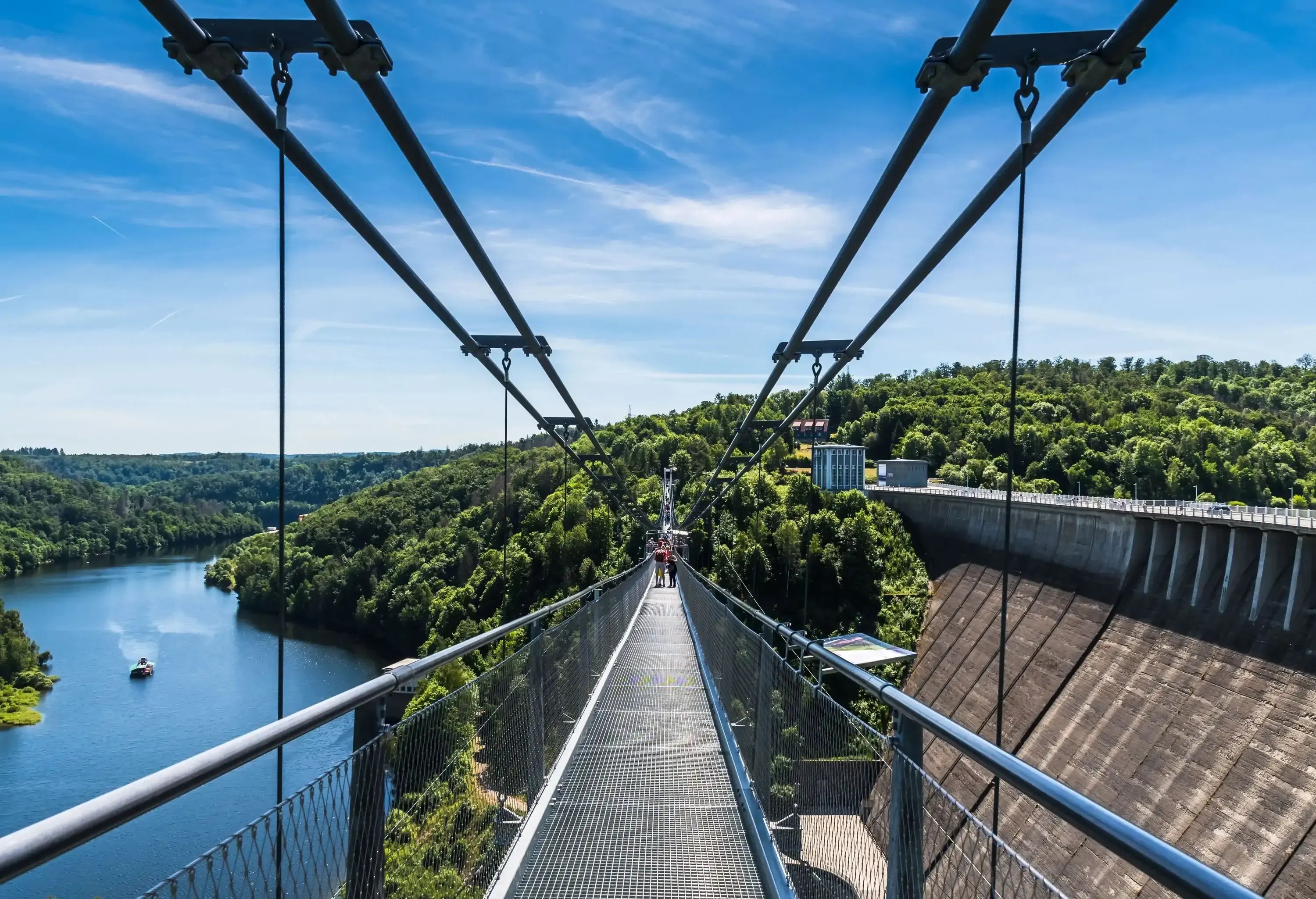 People cross a suspension bridge above a dam with views of a speeding boat in a calm river.