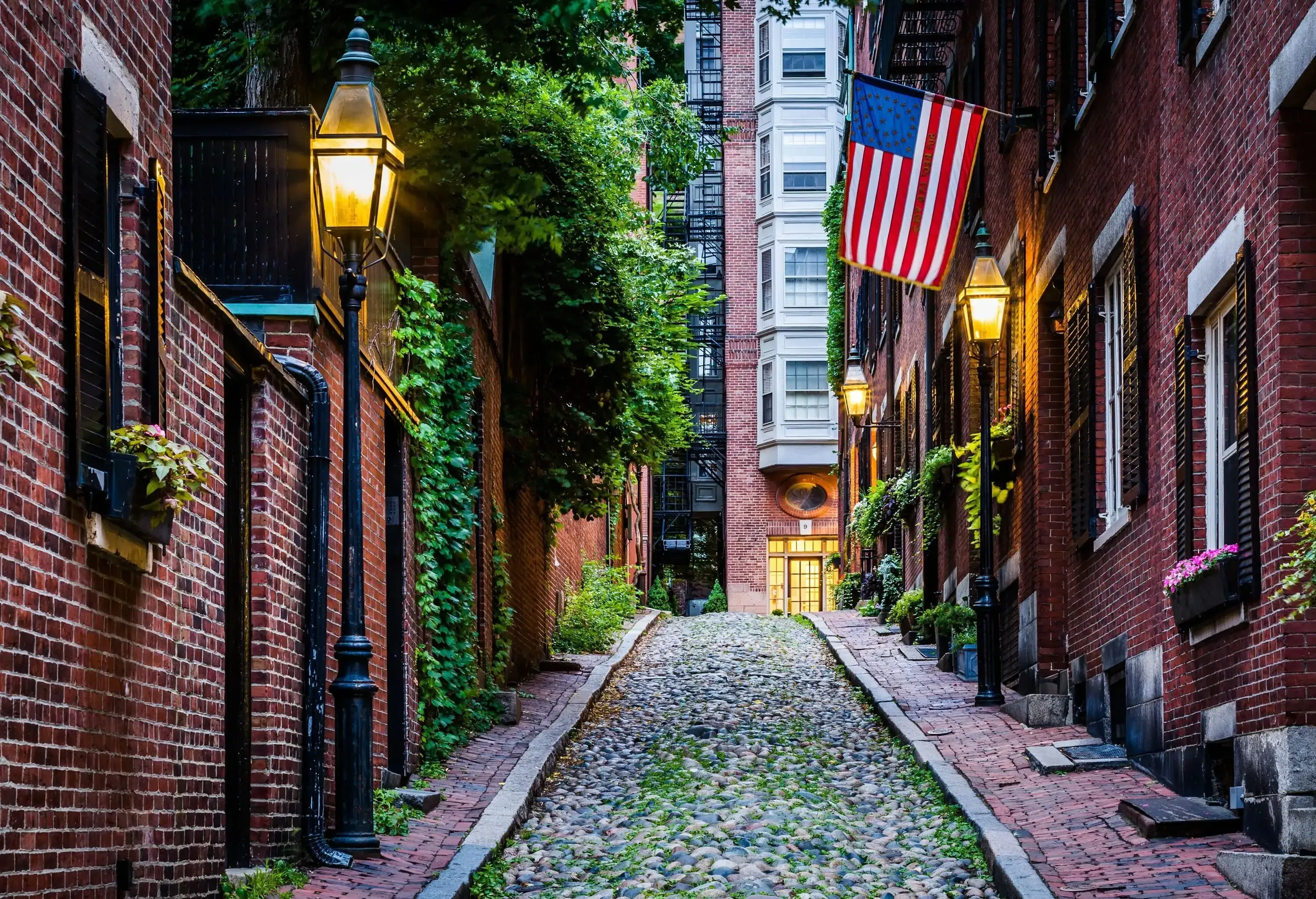 A pebbled street across brick buildings with lush foliage lined with bright streetlamps.