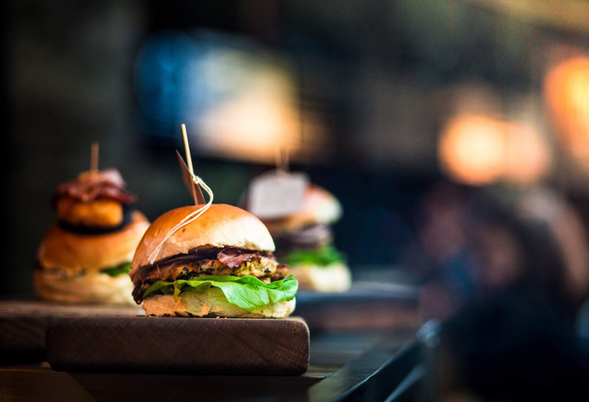 Close up image of a selection of freshly flame grilled burgers in a row on a wooden counter at a London street food market. Each of the burgers has its own label, on which is written the contents of the burger. The burgers are sandwiched between glazed buns, and presented on beds of fresh green lettuce and stuffed with melted cheese and red onion. Horizontal colour image with copy space and beautiful bokeh background.