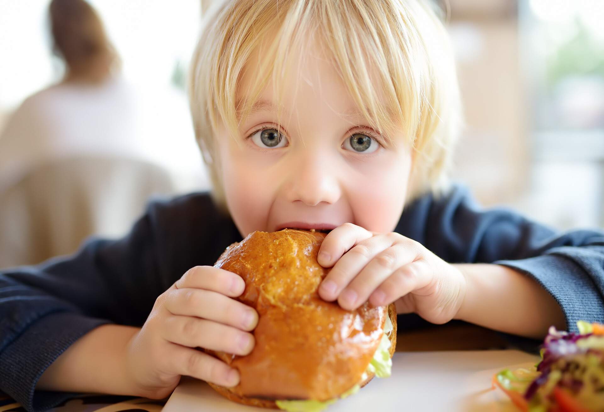 Cute blonde boy eating large hamburger at fast food restaurant. Unhealthy meal for kids. Junk food. Overweight problem child.