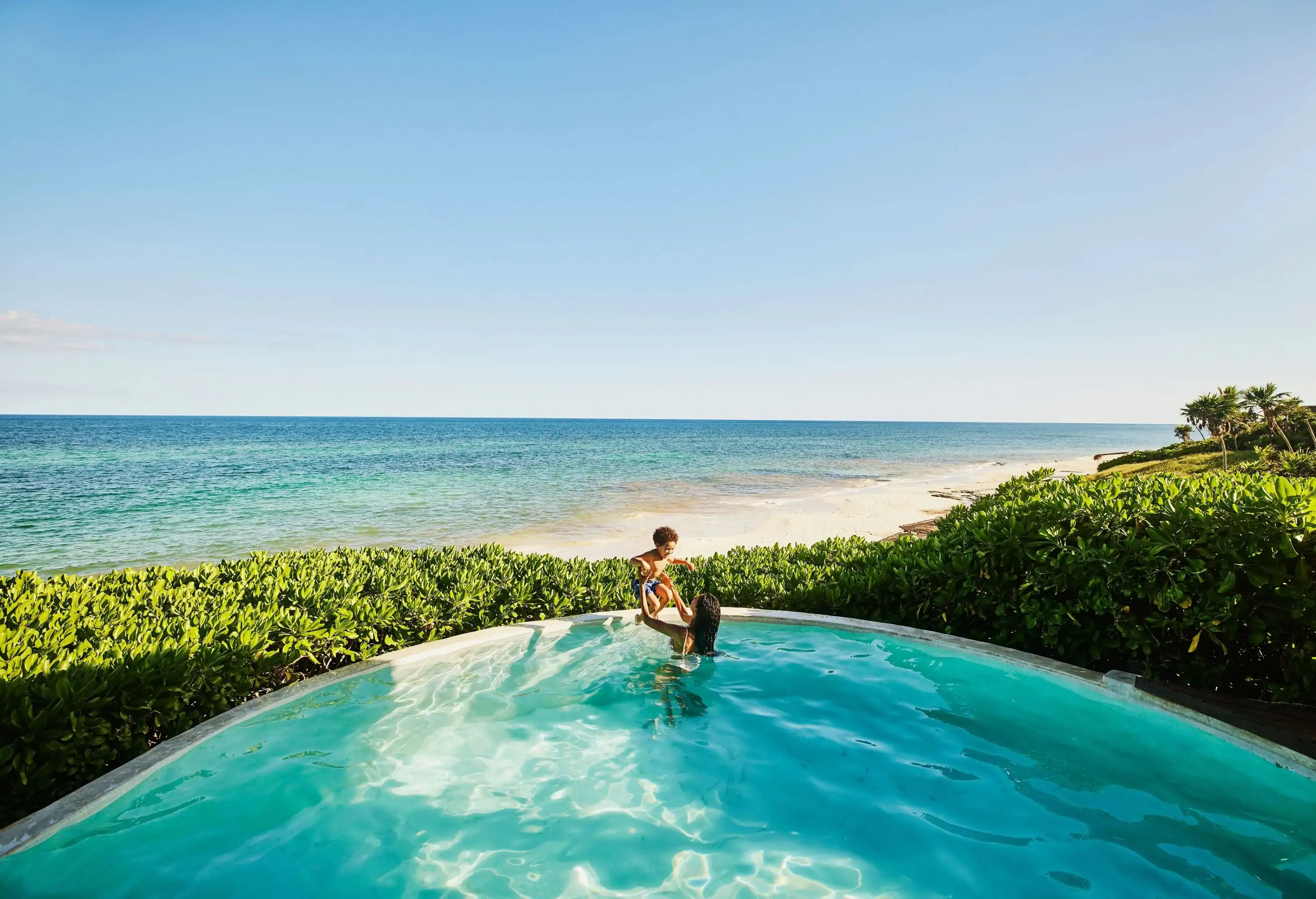 A person lifts a child while immersed in a pool with views of a pristine beach.