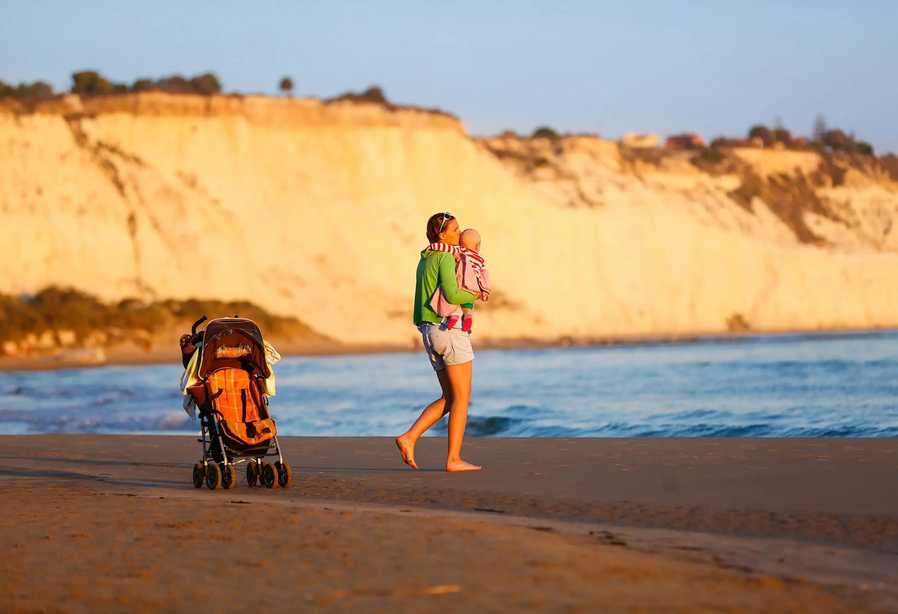 Affectionate mother holding and kissing her baby boy on a sandy beach, on a late summer evening. Family vacation, traveling with children concept.