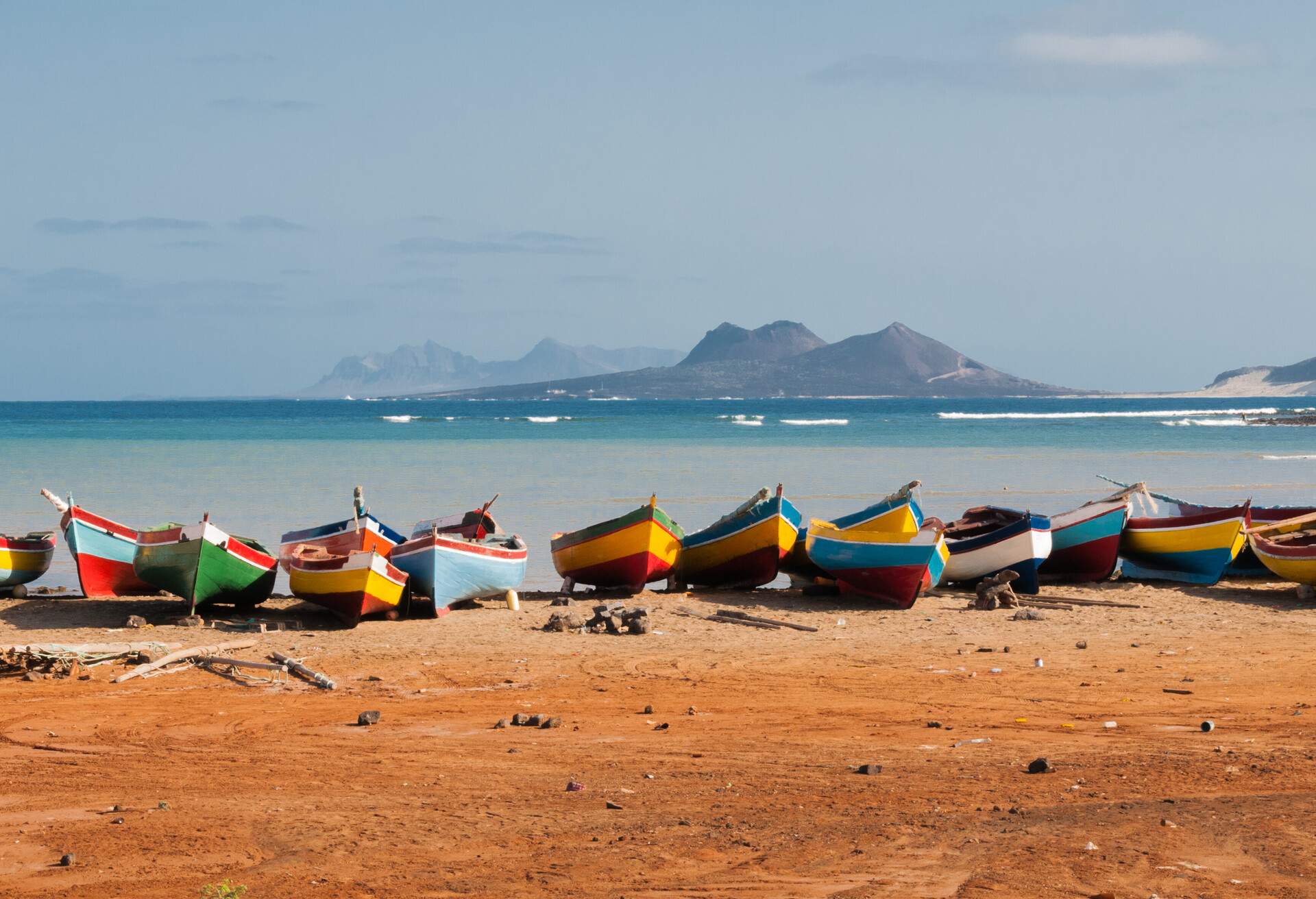 Colourful fishing boats rest peacefully on the sandy beach, their vibrant hues contrasting against the clear blue sea, while majestic mountains rise in the distance.