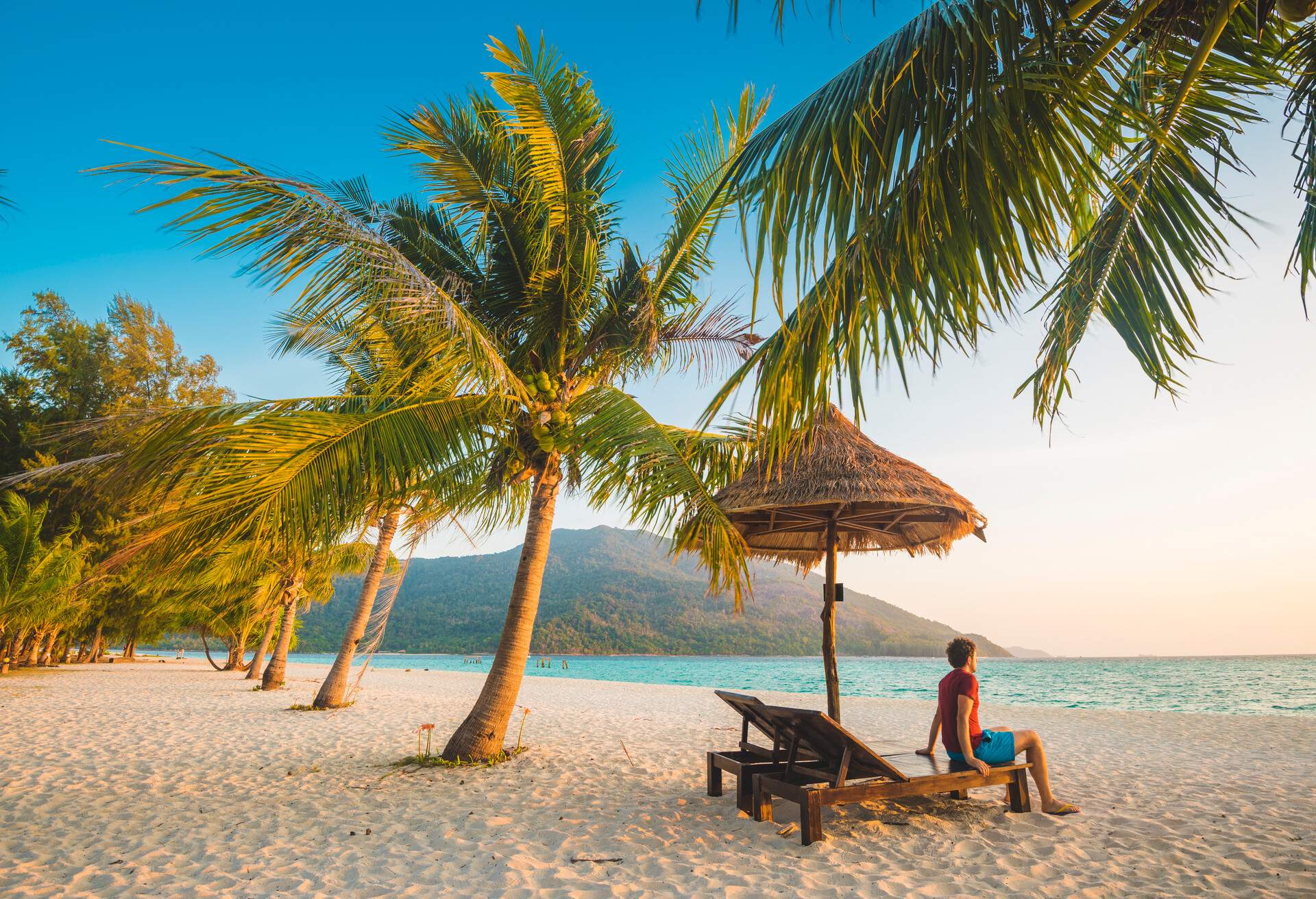 A lone man sits in a wooden sun chair with a straw hut umbrella on the white sand shore by the blue sea at sunrise.