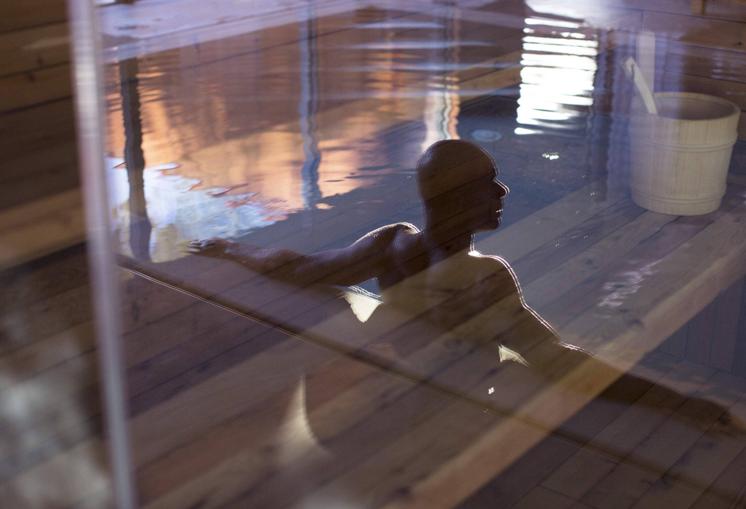 A bald man leans his back on the edge of the pool sauna seen through the glass.