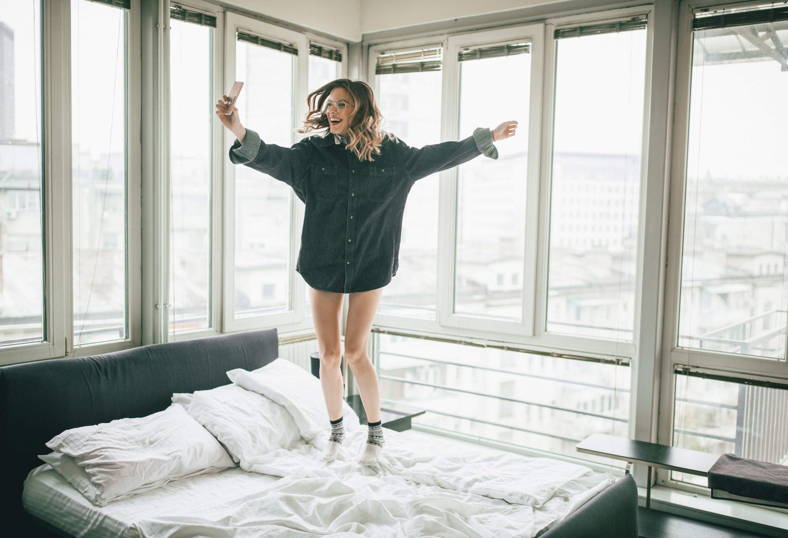 A brunette woman in a denim shirt jumps on the bed of a hotel room as she smiles at her phone.