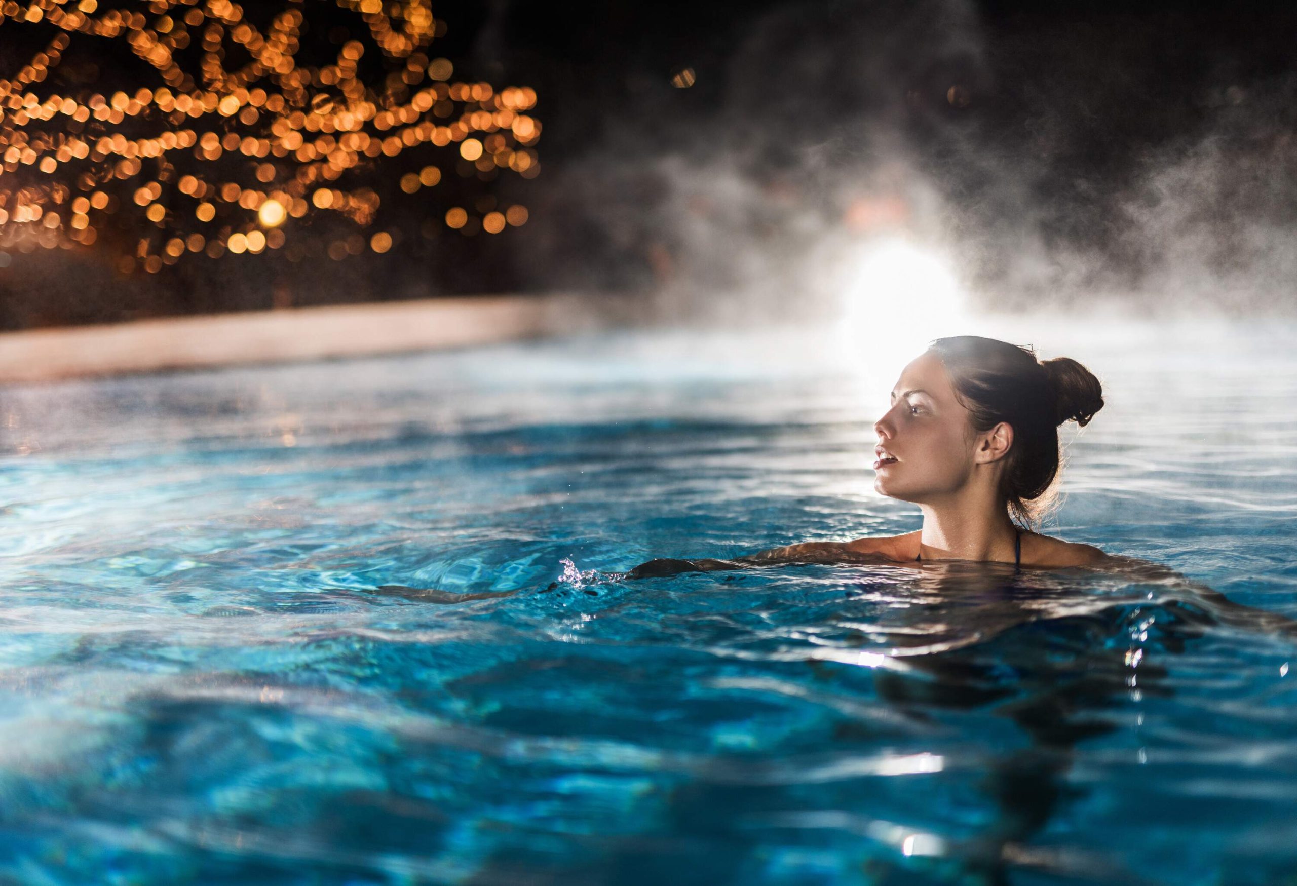 A woman, looking to her right while submerged in a steaming heated swimming pool.