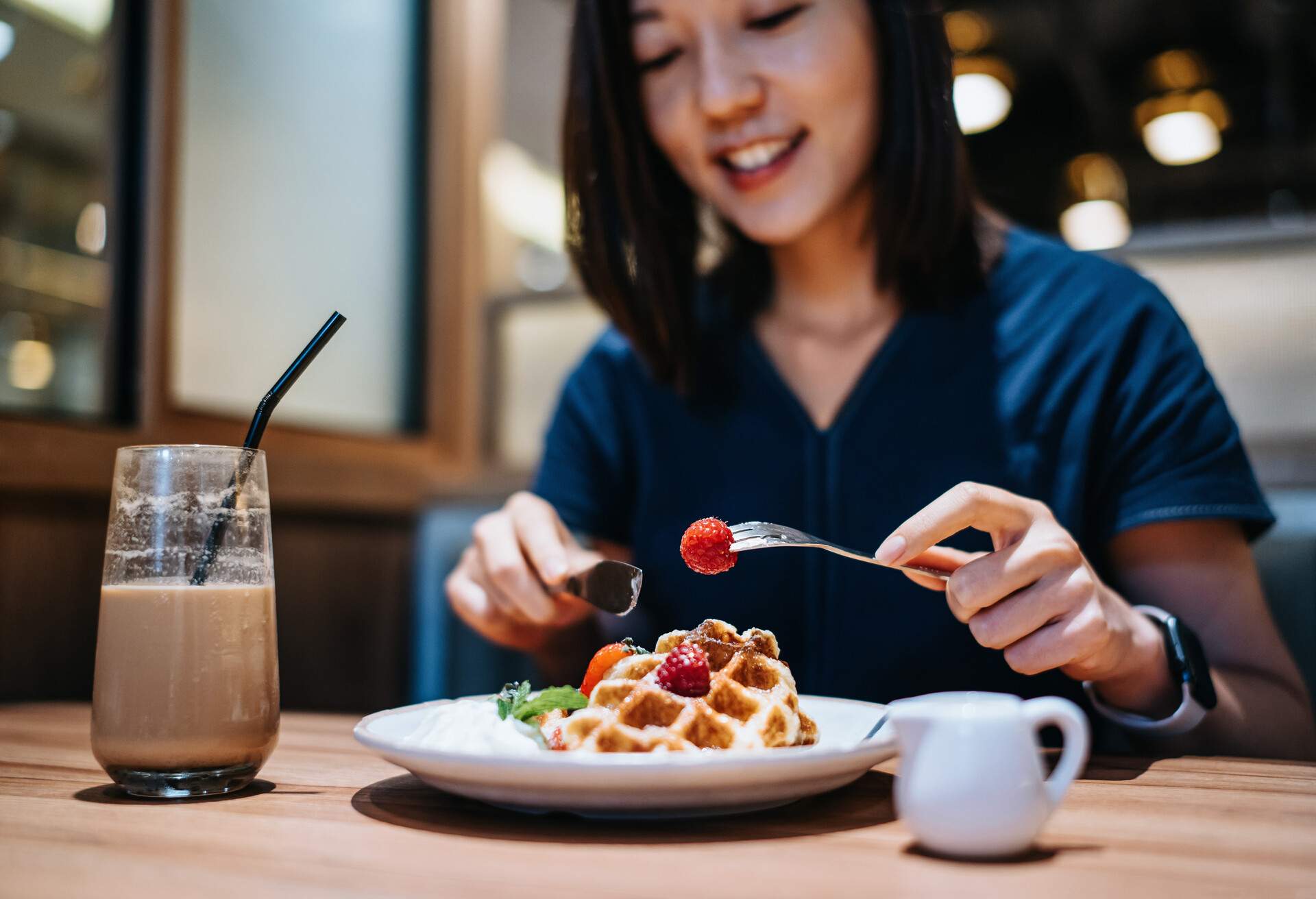 Beautiful smiling young Asian woman eating waffle with fresh cream and strawberries and having coffee in cafe