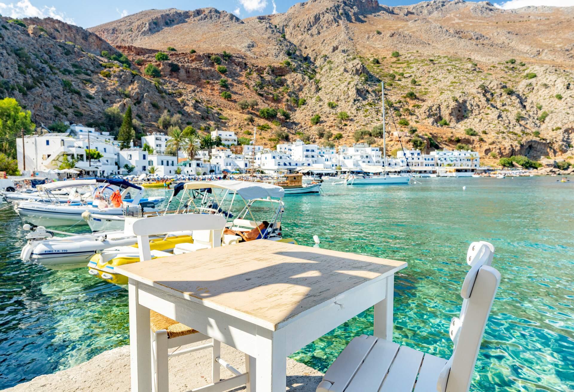 A white table paired with two chairs alongside the docked boats on a tidy harbour in a white-washed village at the foot of the mountain.