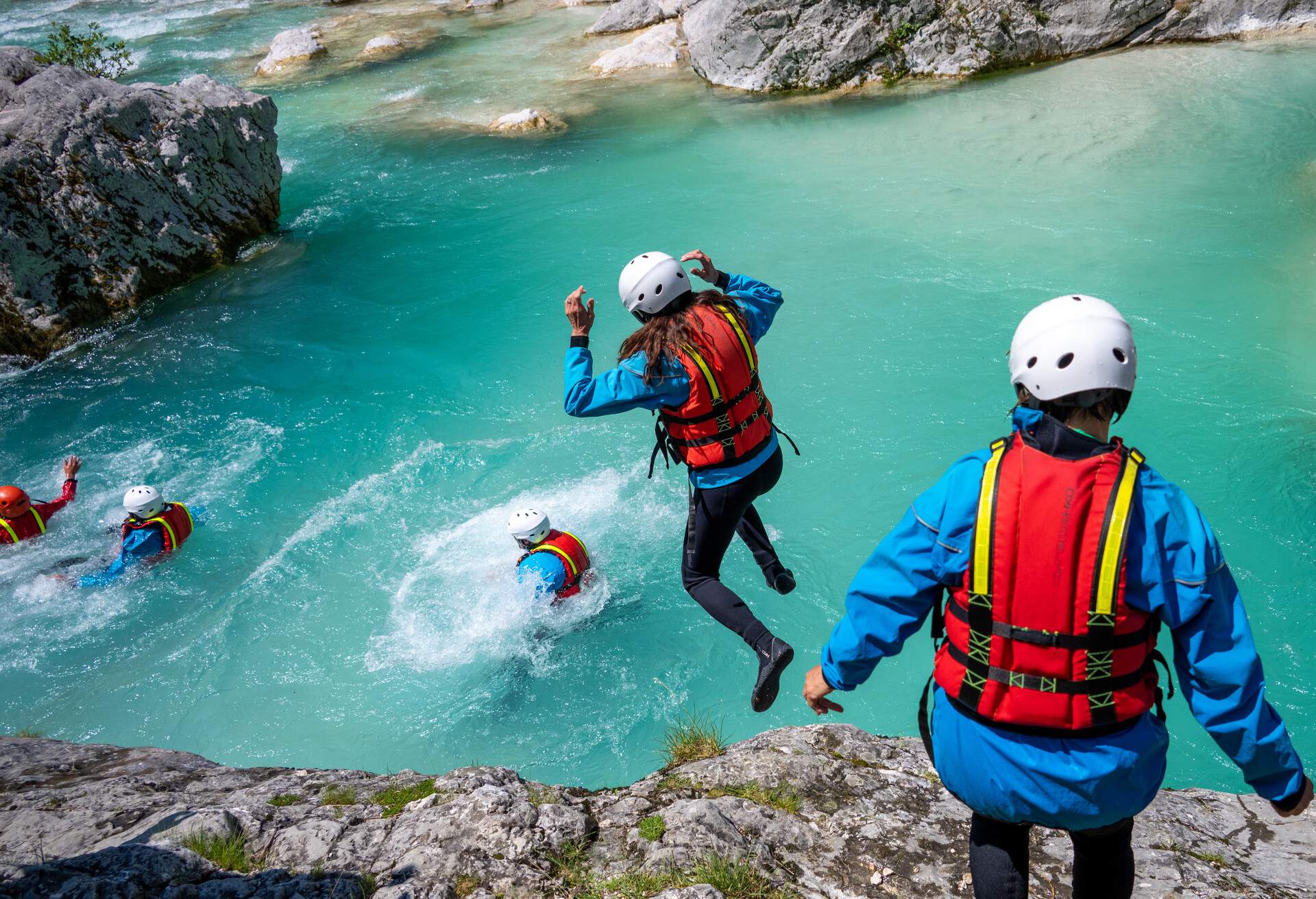 People in canyoning gear jumping one by one off a ledge into a river.