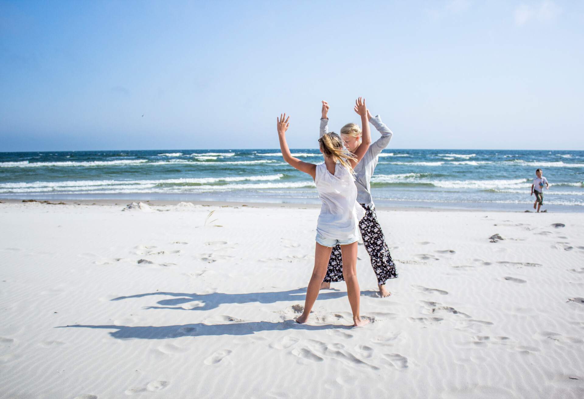 Two young girls pose with their arms up on the sand, with a little boy running to the shore in the background, Skane, Sweden